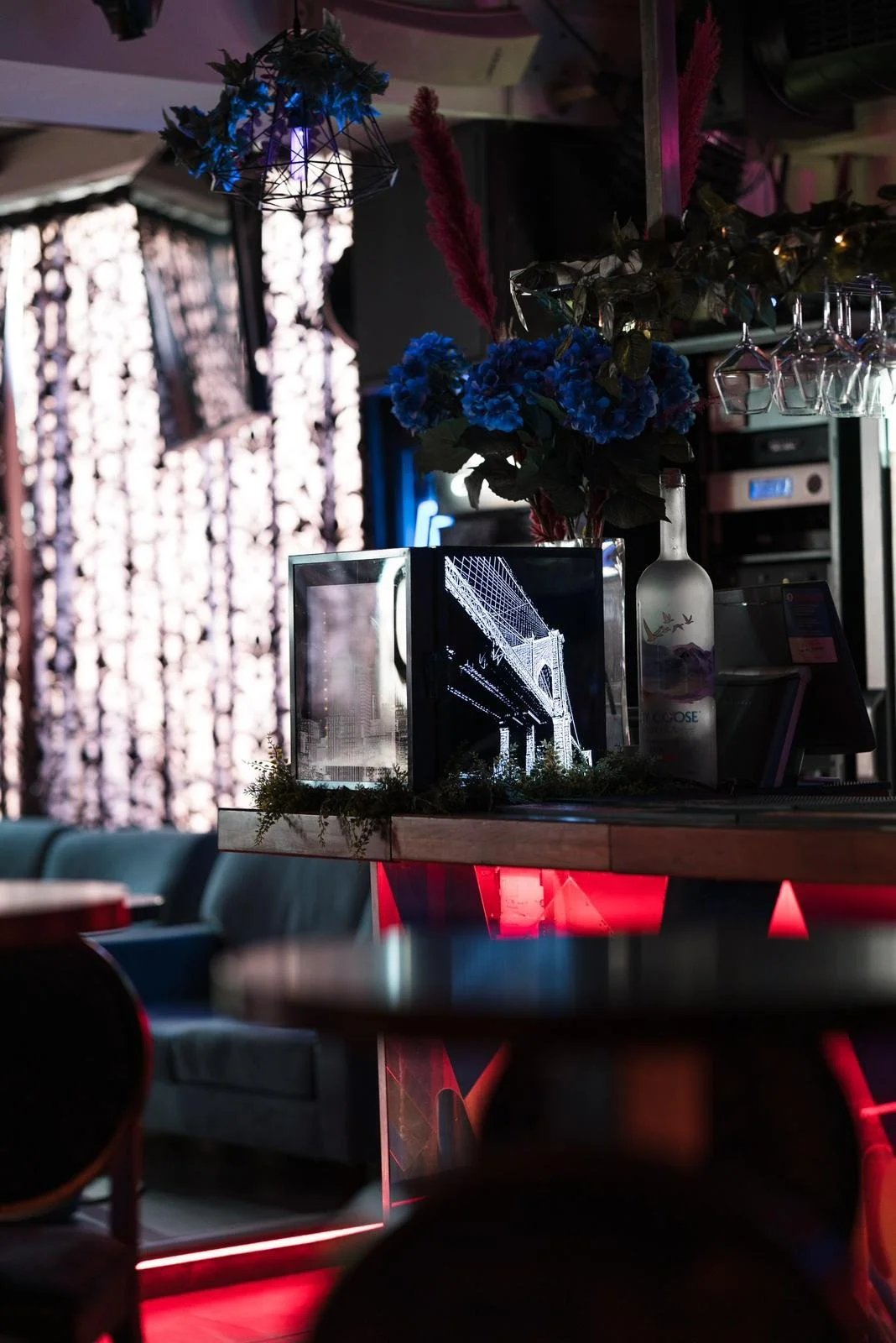 Interior of a dimly lit bar with decorative flowers, including blue and pink blossoms, a neon light, and an electronic display showing the Brooklyn Bridge.