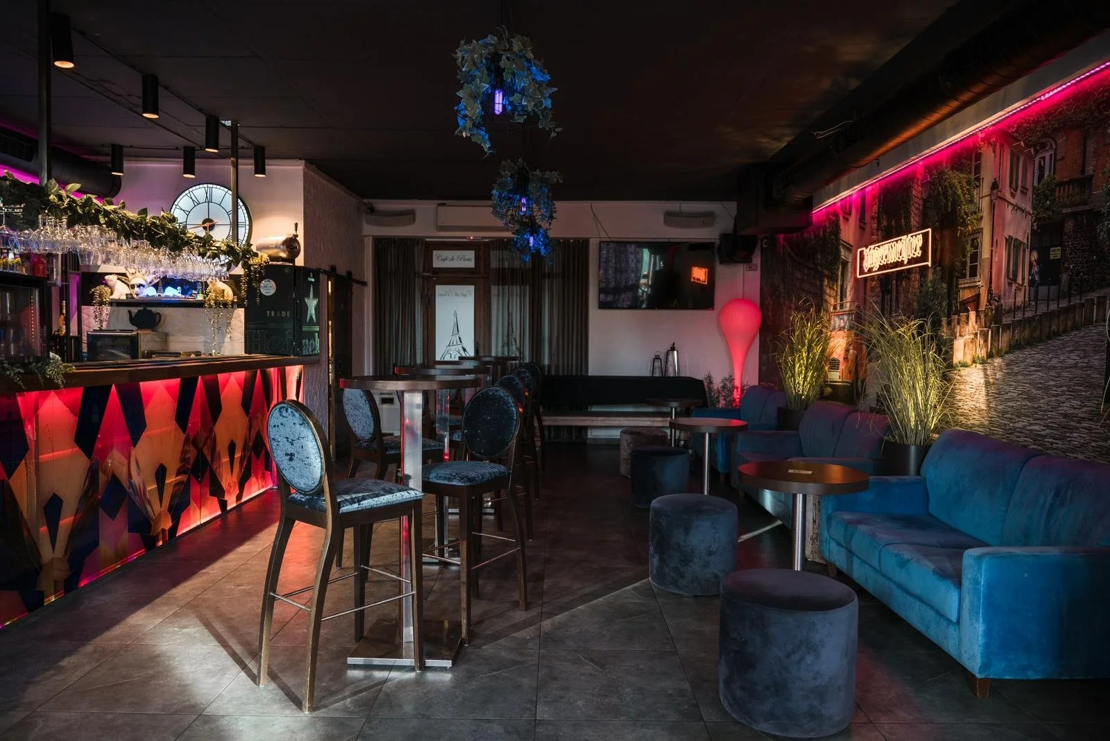 Interior of a dimly lit bar or lounge with colorful neon lights, a bar counter with high chairs, and various seating options including a blue sofa and small round stools. Decorative plants and wall art are visible.