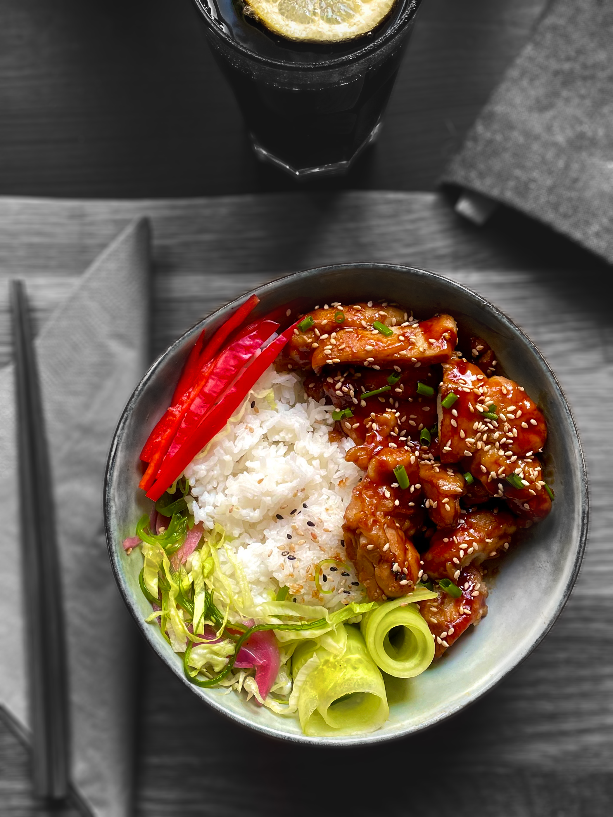 En skål med asiatisk mat innehållande friterad kyckling i sås, ris och grönsaker som skivade röda paprikor, grön sallad och salladslök. Skålen står på ett träbord tillsammans med ett par ätpinnar och ett glas med mörk läsk med en citronskiva i. 