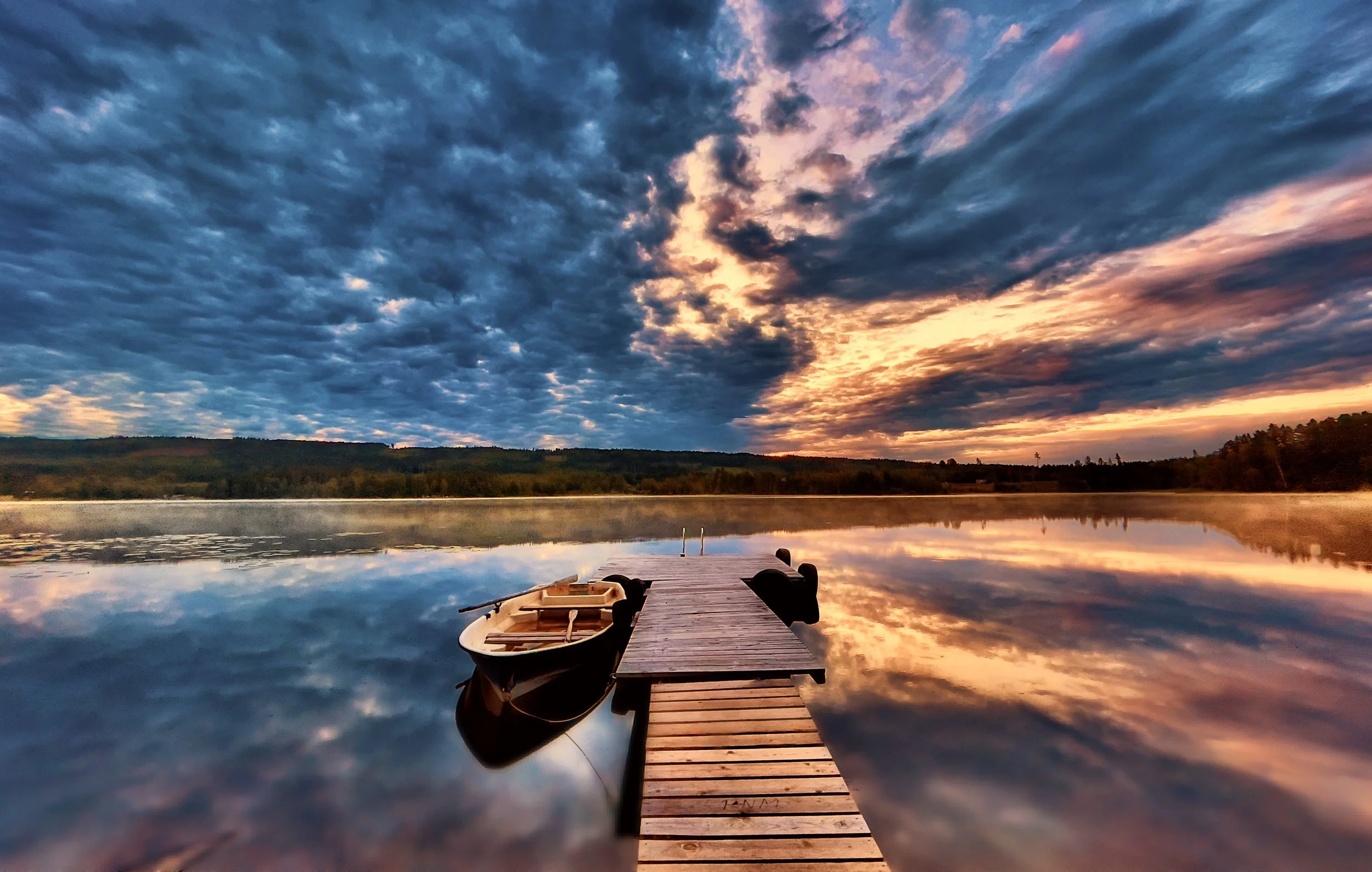 En brygga som går ut över en lugn sjö i solnedgång. Vid bryggan finns en roddbåt fastsnurrad. Vattnet i sjön speglar en dramatisk himmel i bildens överkant. 