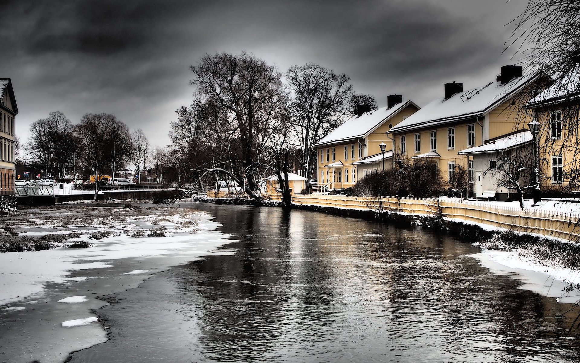En bild tagen på vintern med en å med vatten som nästan är fruset, snö bredvid ån och gula hus vid sidan om ån. Svartvit himmel och mörka kala träd. 