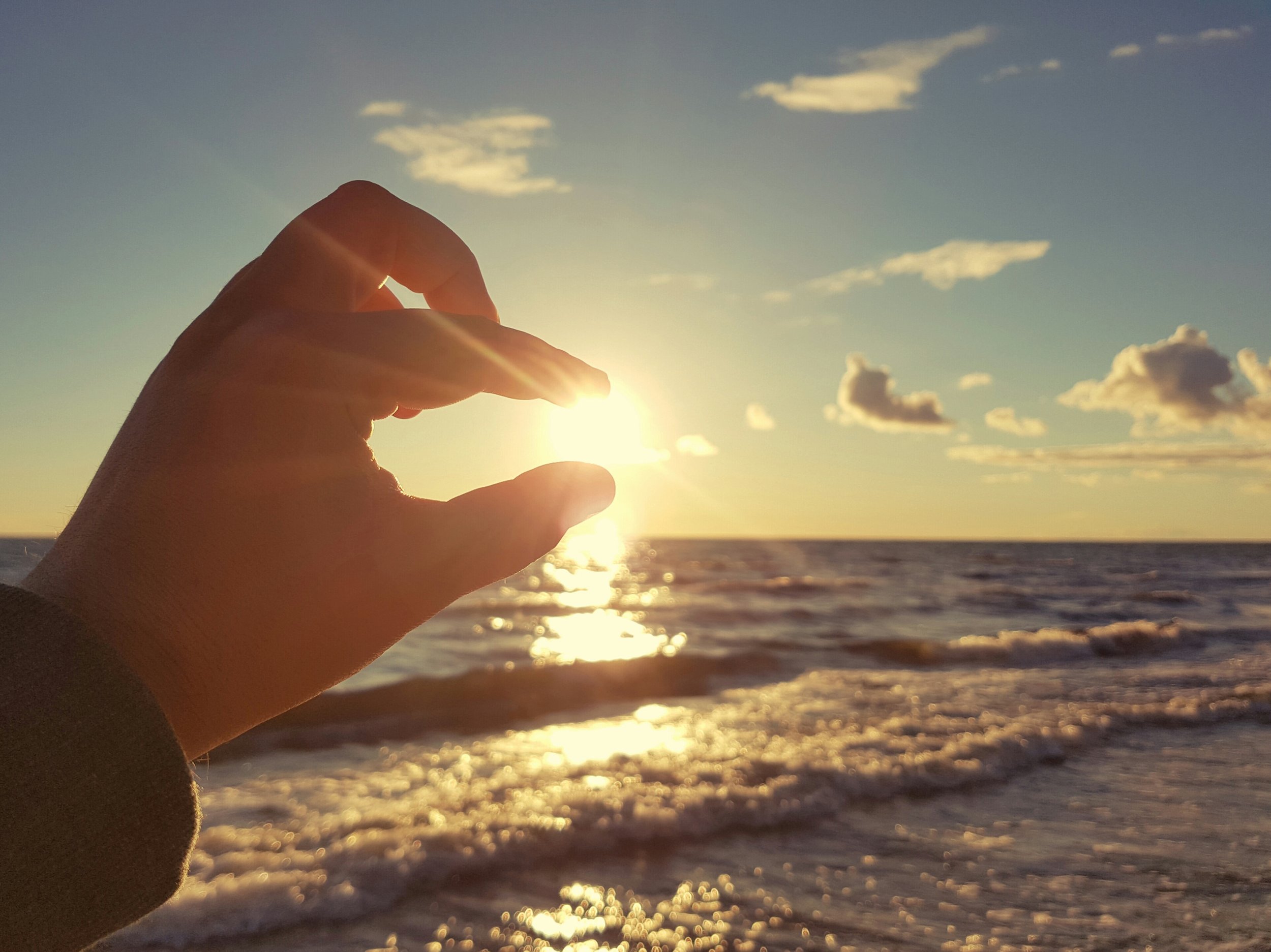 En hand som med fingrarna håller solen. Bilden är tagen på en strand med havet i bakgrunden. 
