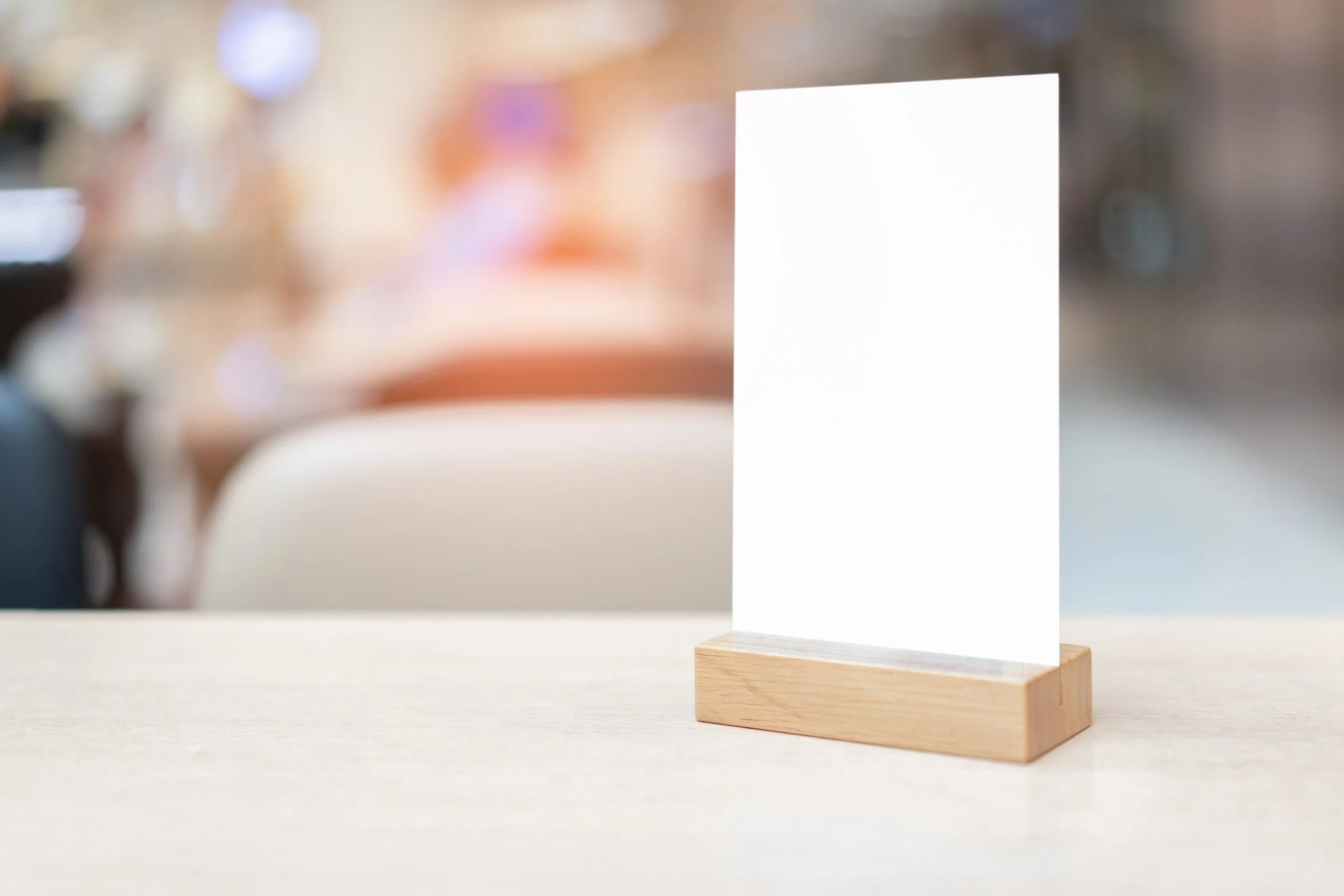 A blank white sign with a wooden base on a light-colored tabletop in a blurred background of a restaurant or cafe.