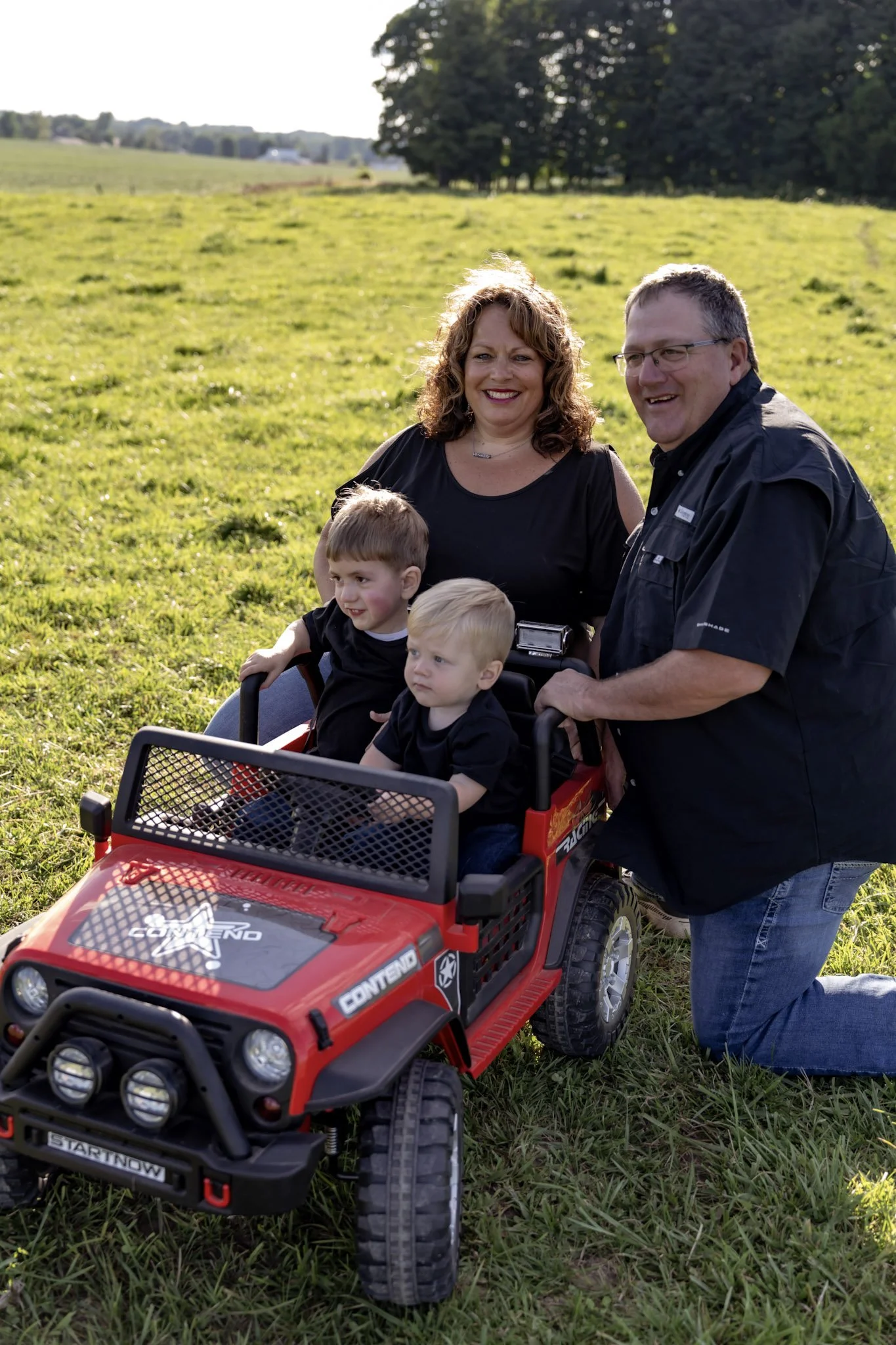 Two of Julie's grandchildren sitting in a toy red truck with Julie and her husband on a grassy field during daytime, with trees in the background.