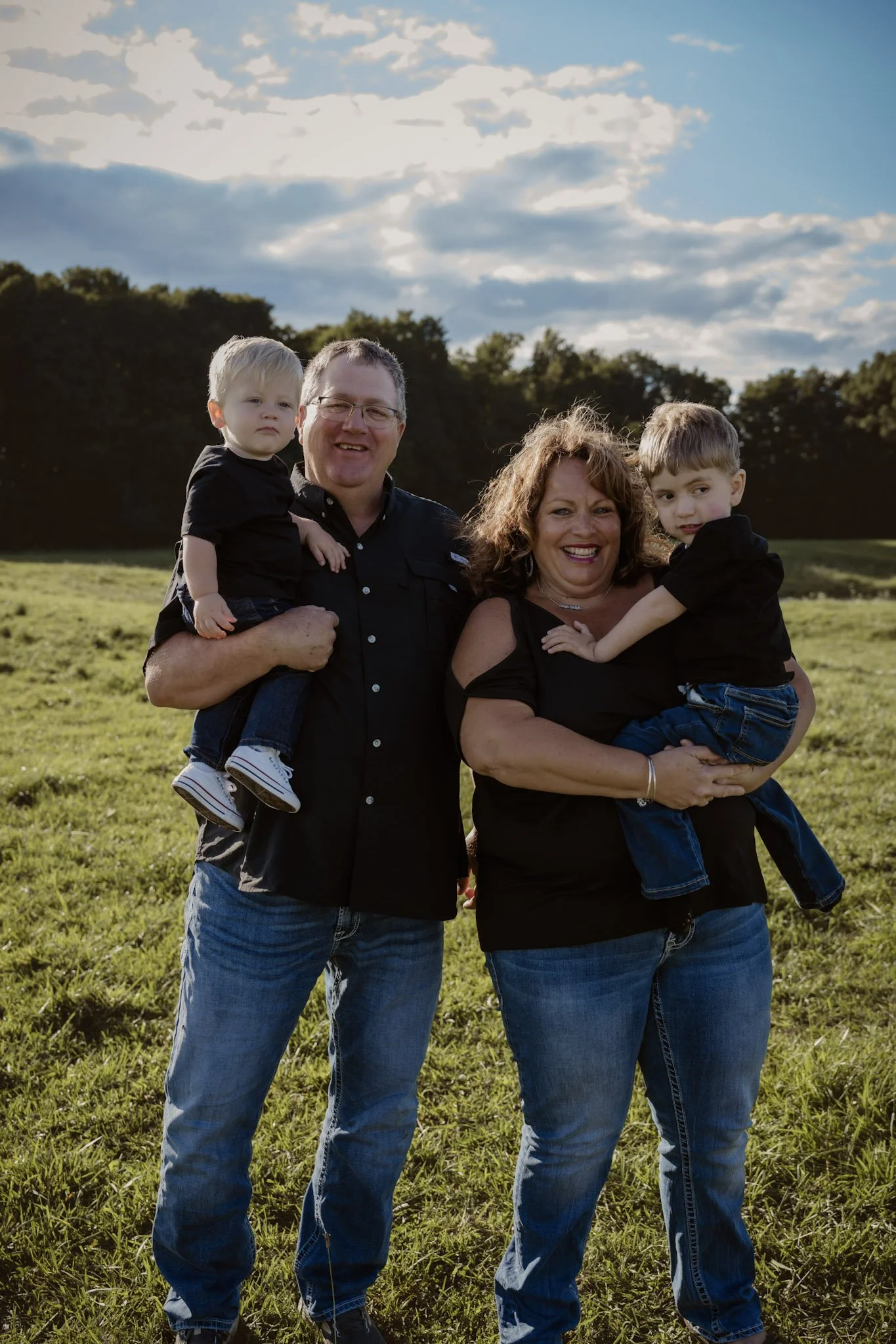 A family of four outdoors on a grassy field, with two parents and two young boys, all smiling and dressed in casual dark clothing, against a background of trees and a partly cloudy sky.