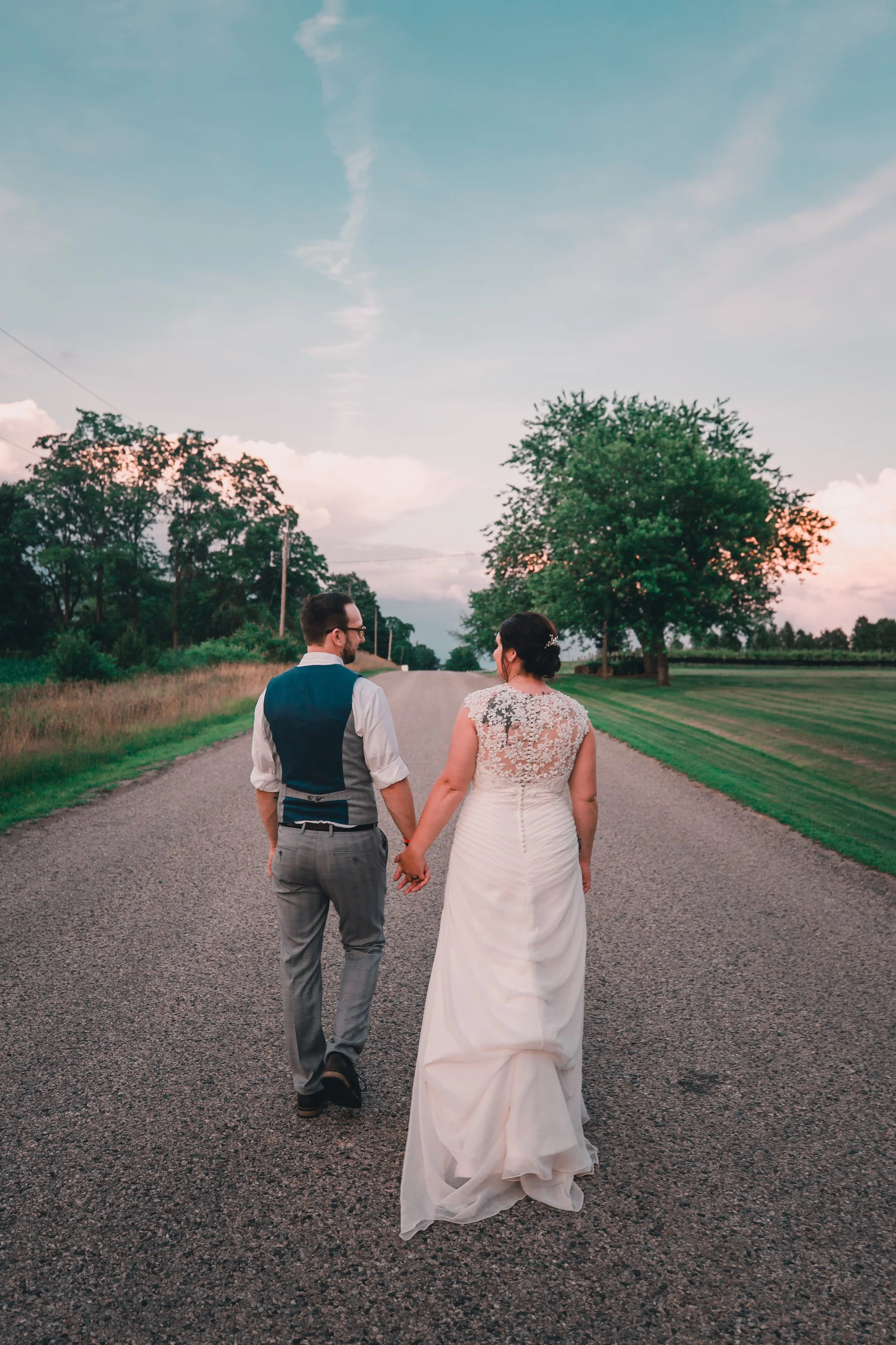 A couple holds hands and walks down a rural road at sunset. The woman wears a white wedding dress with lace details, and the man wears a white shirt with rolled sleeves, a blue vest, and gray pants. Tall trees line the road amid a green landscape.