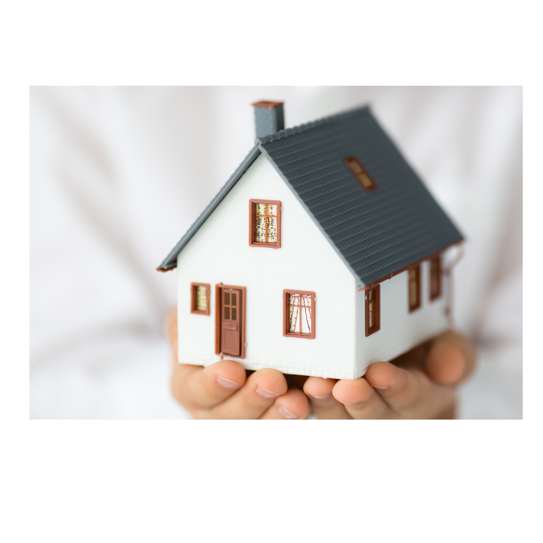Person holding a small model house with white walls, brown window frames, a brown door, a gray roof, and a chimney.