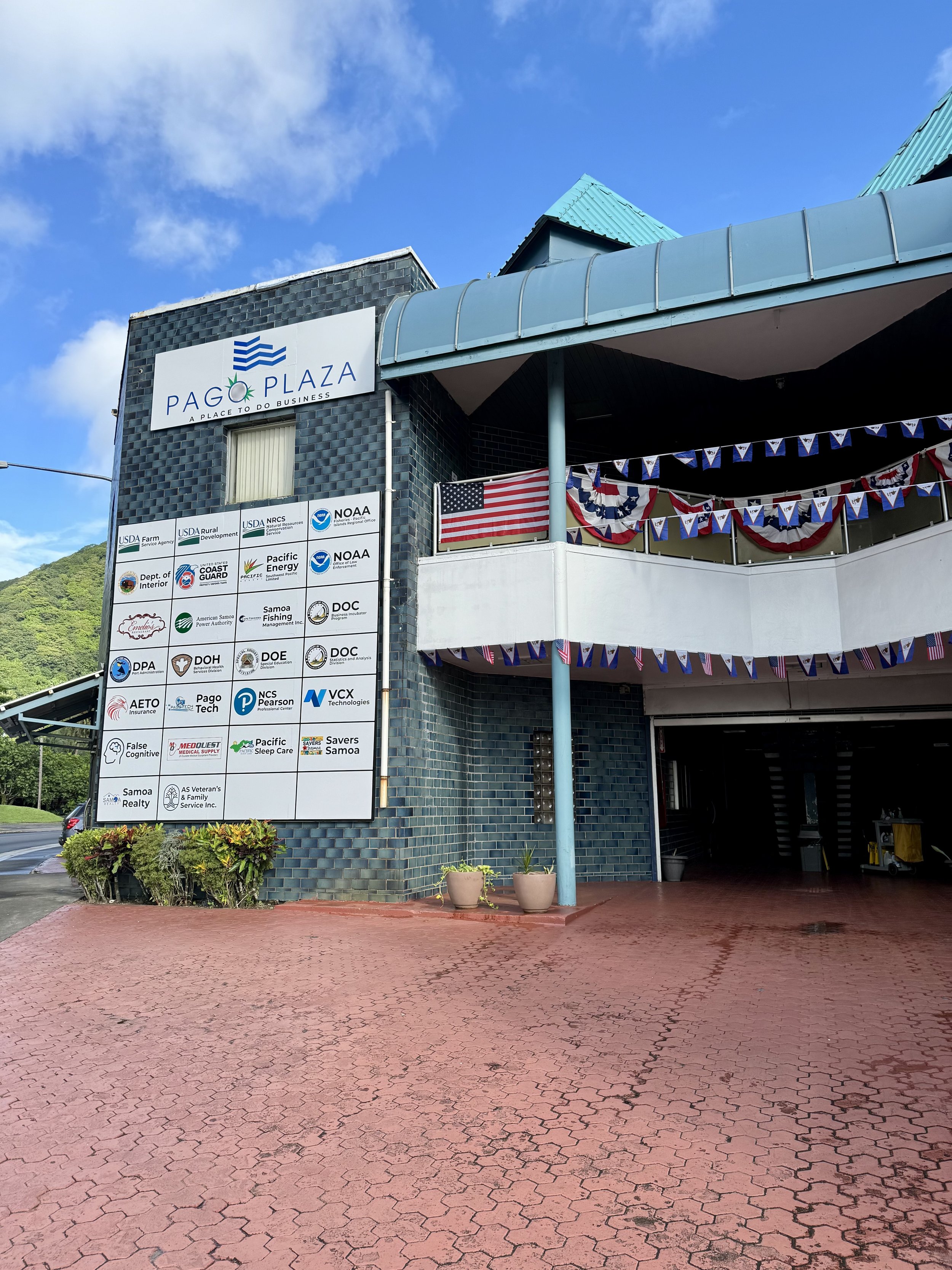 Exterior of Pago Plaza with a sign displaying various business logos, patriotic bunting, an American flag, and potted plants outside on a sunny day.