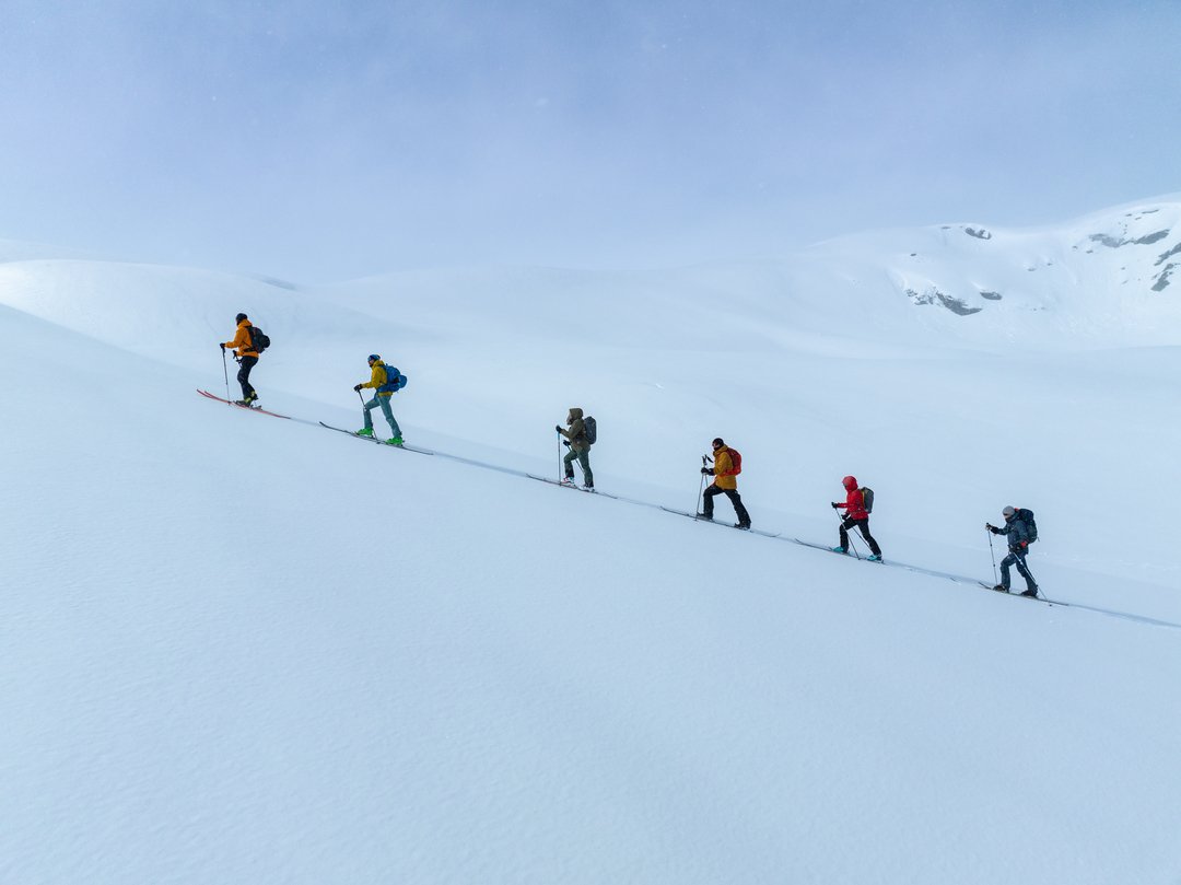 Seks personer på ski opp en snøtopp med snødekte fjell i bakgrunnen.