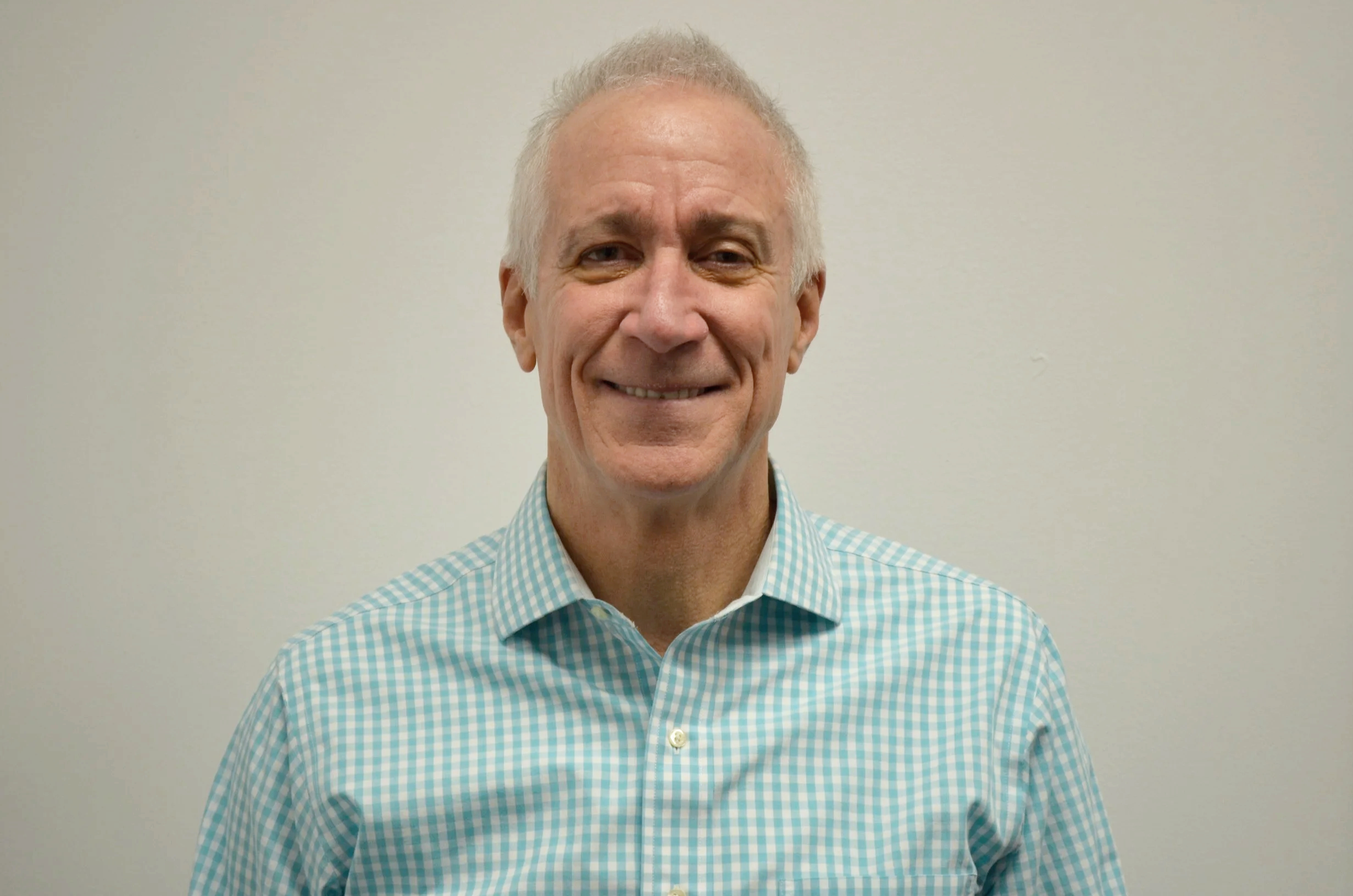A smiling middle-aged man with gray hair and a checkered button-up shirt standing against a plain light-colored wall.