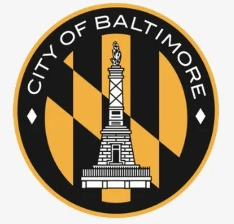 The emblem of the city of Baltimore featuring a statue on a pedestal with black and gold colors and the words 'City of Baltimore' around it.