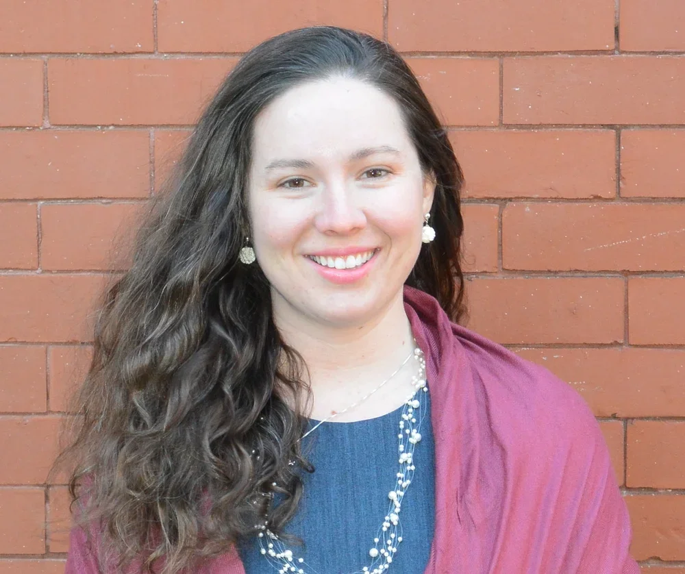 A woman with long, curly brown hair smiling at the camera, standing against a red brick wall. She is wearing a dark blue top, a burgundy shawl, and pearl jewelry.
