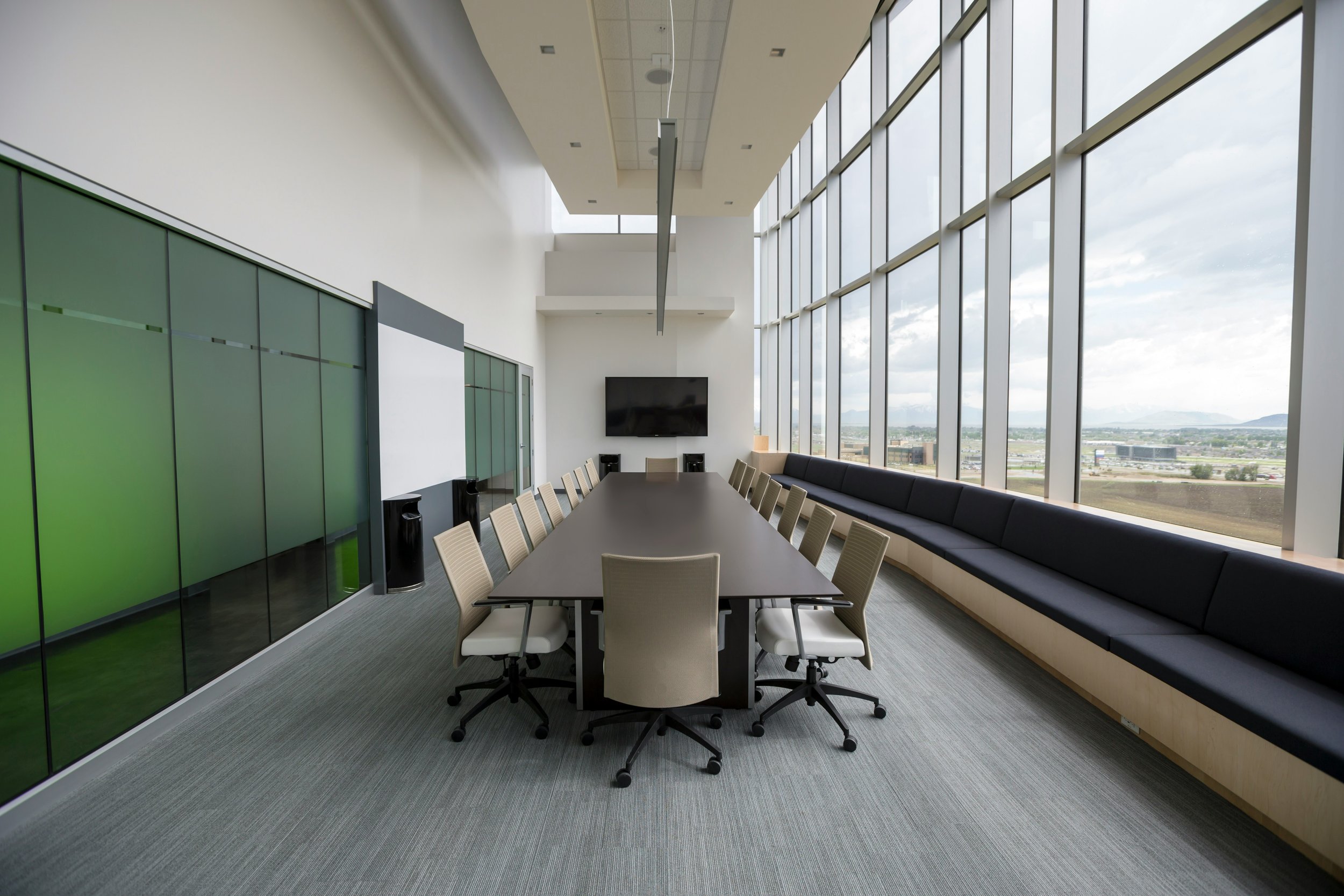 Modern conference room with a long table surrounded by beige office chairs, large windows with city view, wall-mounted television, and a black built-in bench along the window, with a gray carpeted floor.
