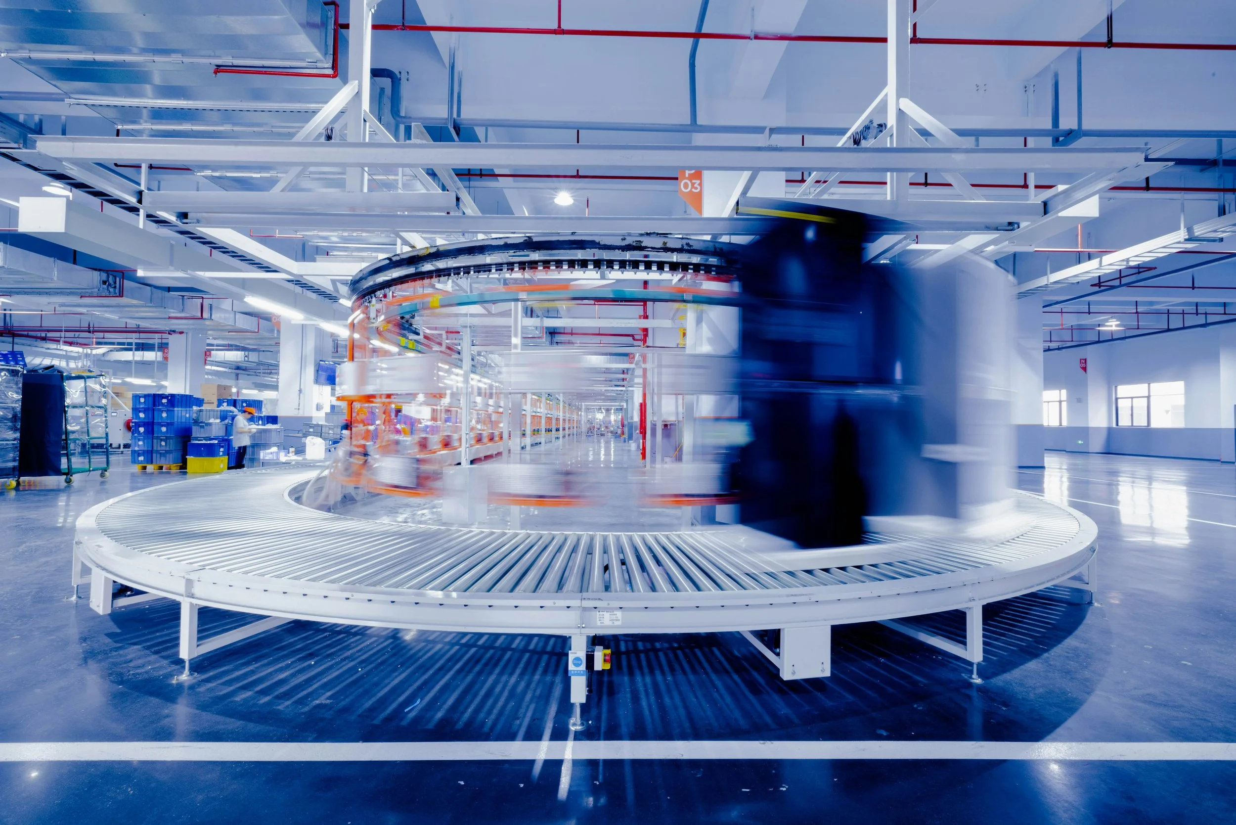 Motion-blurred robotic conveyor belt in a modern, brightly lit warehouse or manufacturing facility with white and blue color scheme.