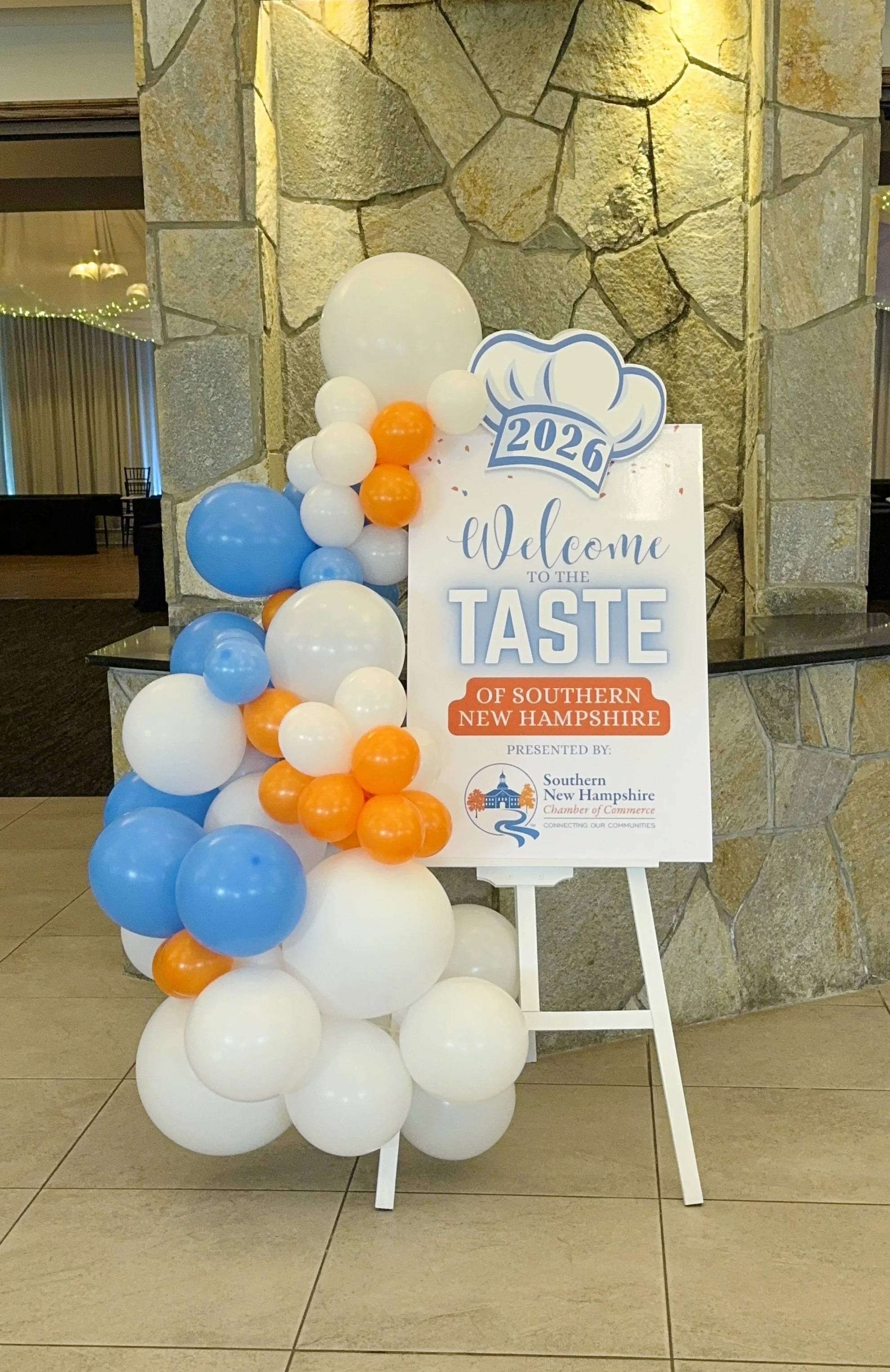 Entry easel balloon arrangement featuring white, blue, and orange balloons with custom sign welcoming guests to the Taste of Southern New Hampshire 2026 event, presented by Southern New Hampshire Chamber of Commerce at Castleton Banquet Venue Windham