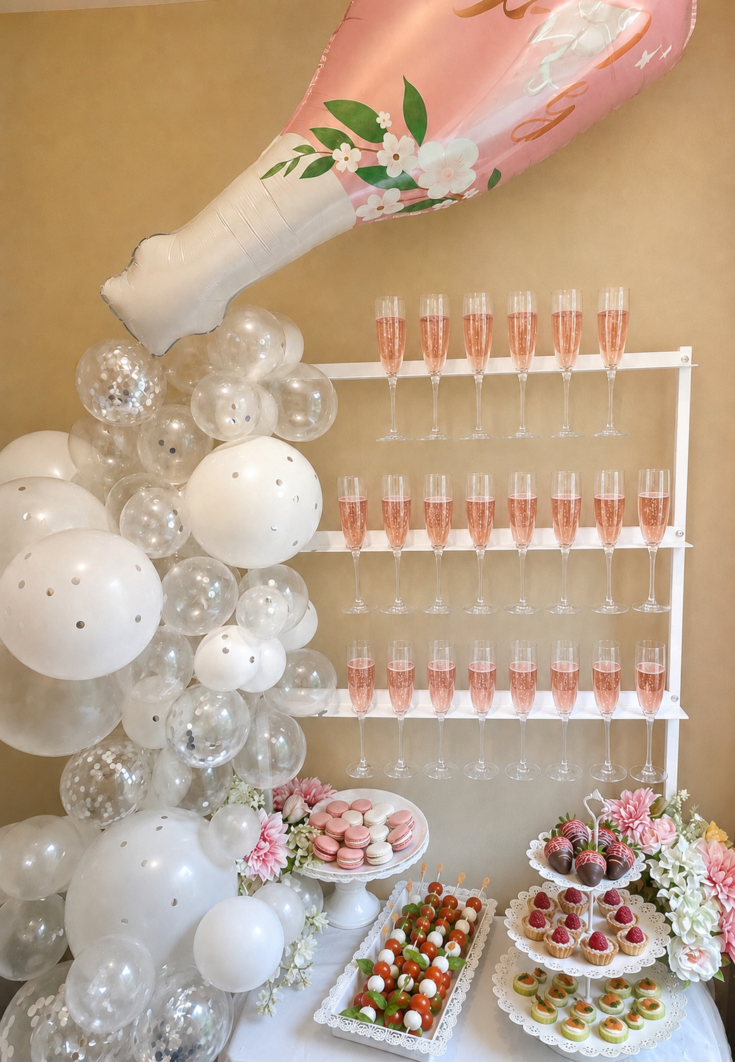 A white Champagne serving rack shelf with a large pink foil balloon shaped like a champagne bottle and smaller balloons glasses of pink champagne in flutes presented on shelves with assorted desserts and decorative flowers.