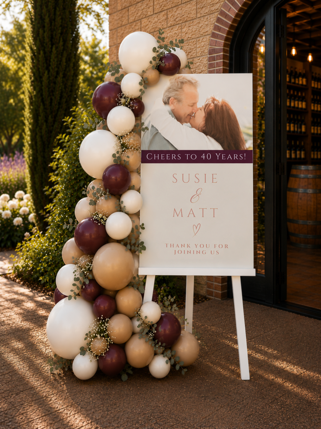 40th anniversary party welcome sign with a couple's photo, decorated with a beige, white and burgundy balloon garland and greenery outside a winery venue.