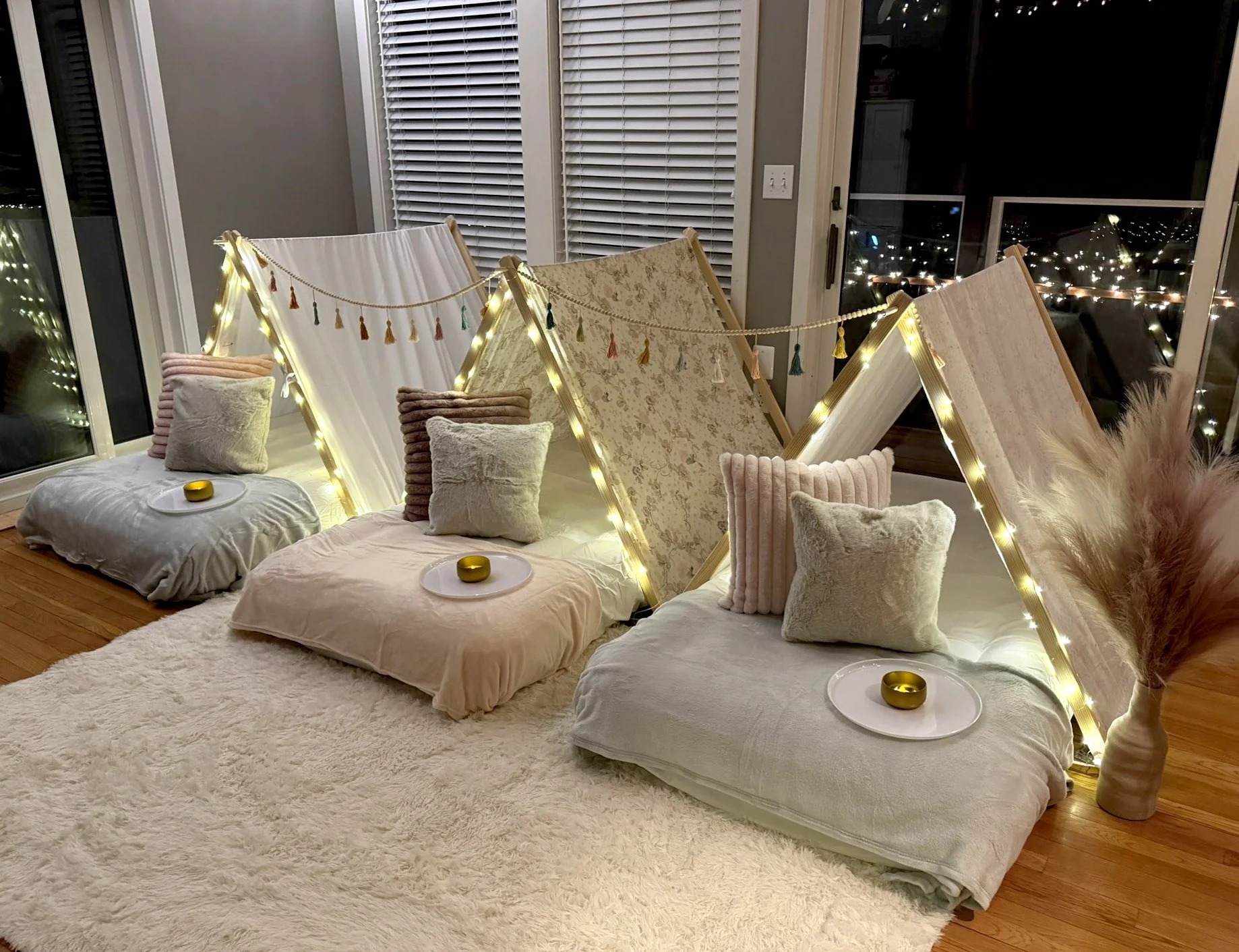Three indoor sleepover tents on the floor with airbeds, pillows and blankets, set up for a cozy glamping sleepover party. Each teepee area has a white plate with a small, gold container, and illuminated by string lights.