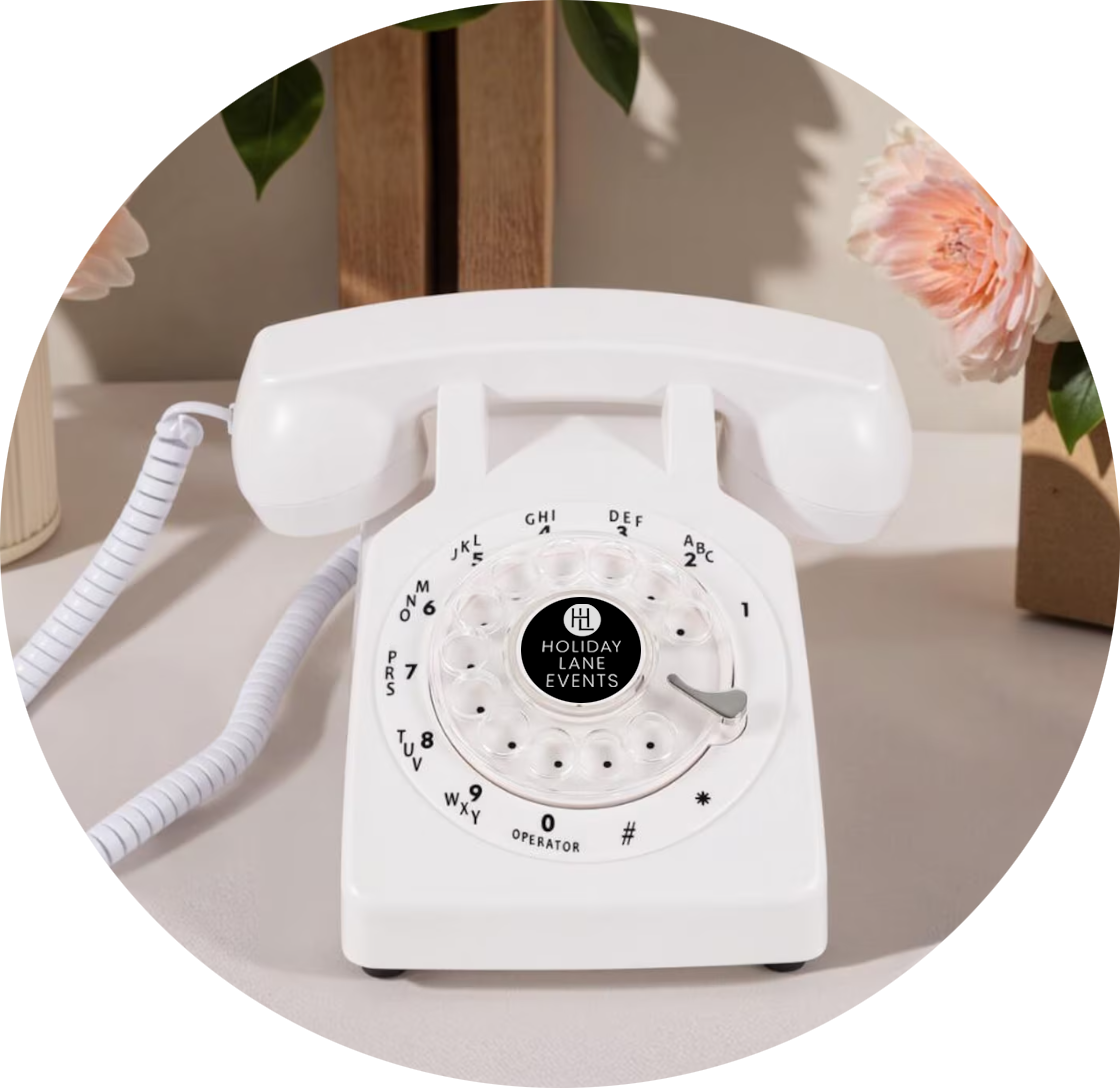 White vintage rotary telephone with a black center displaying 'Holiday Lane Events', placed on a table with pink flowers and green leaves in the background.
