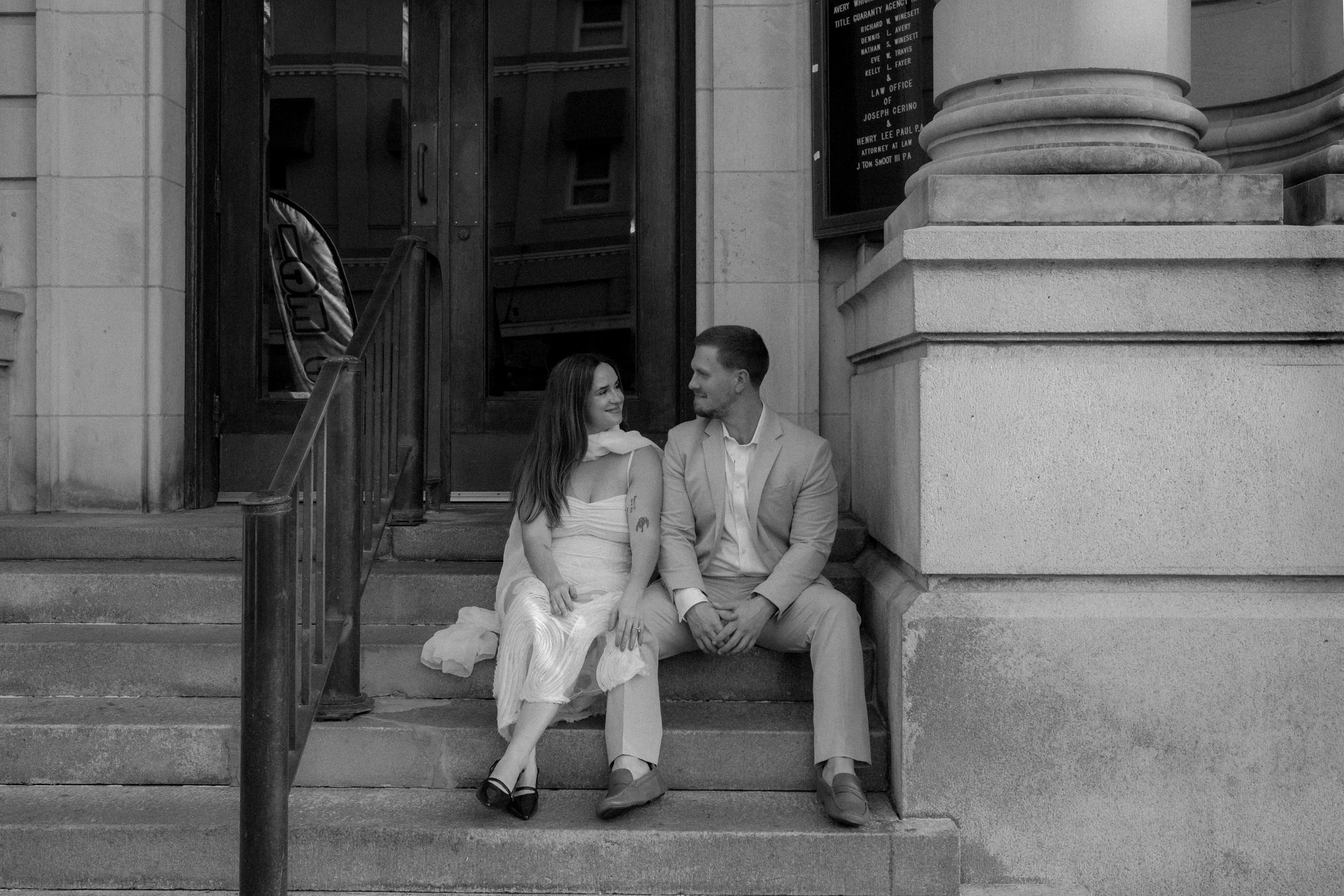 A black and white photo of a man and woman sitting on steps outside a building, looking at each other and smiling. The woman wears a light dress and has long hair, while the man is dressed in a suit with a light blazer and trousers.