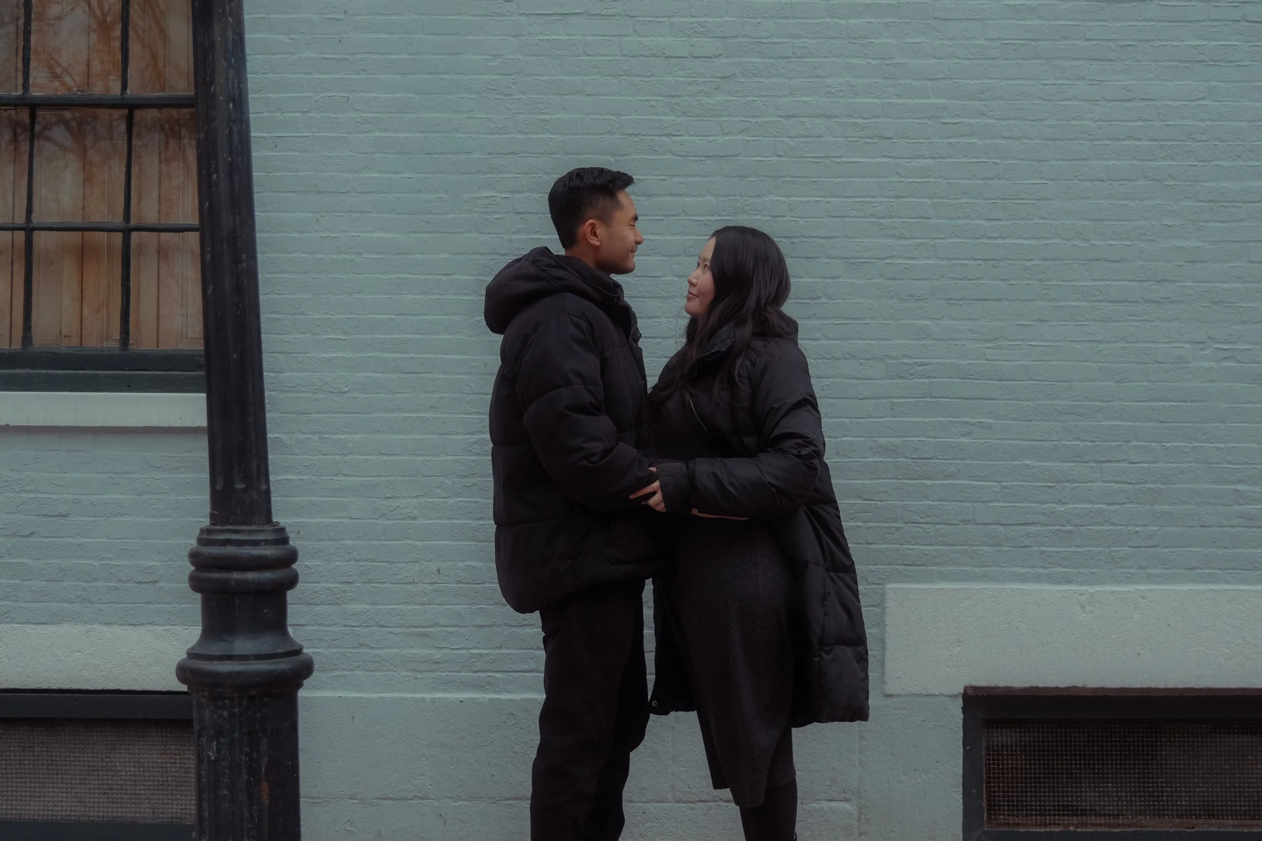 A man and woman in warm clothing facing each other and smiling, standing close against a light blue brick wall beside a black lamppost in an urban setting.