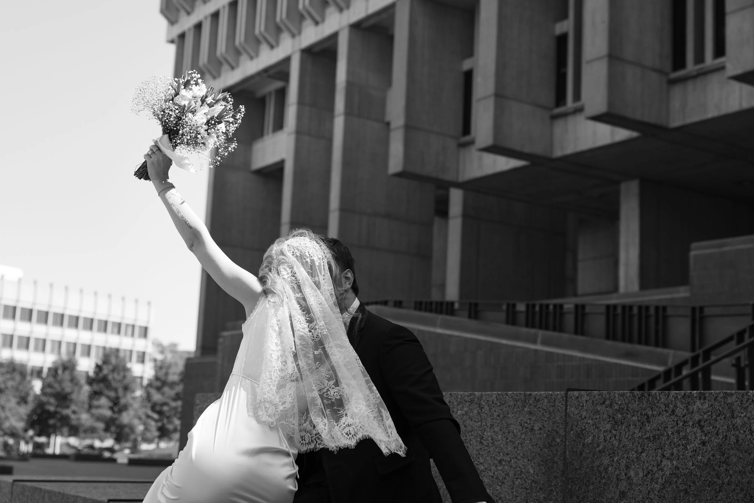 A black and white photograph of a bride holding a bouquet of flowers high in the air, standing next to a groom in an urban setting with modern buildings in the background.