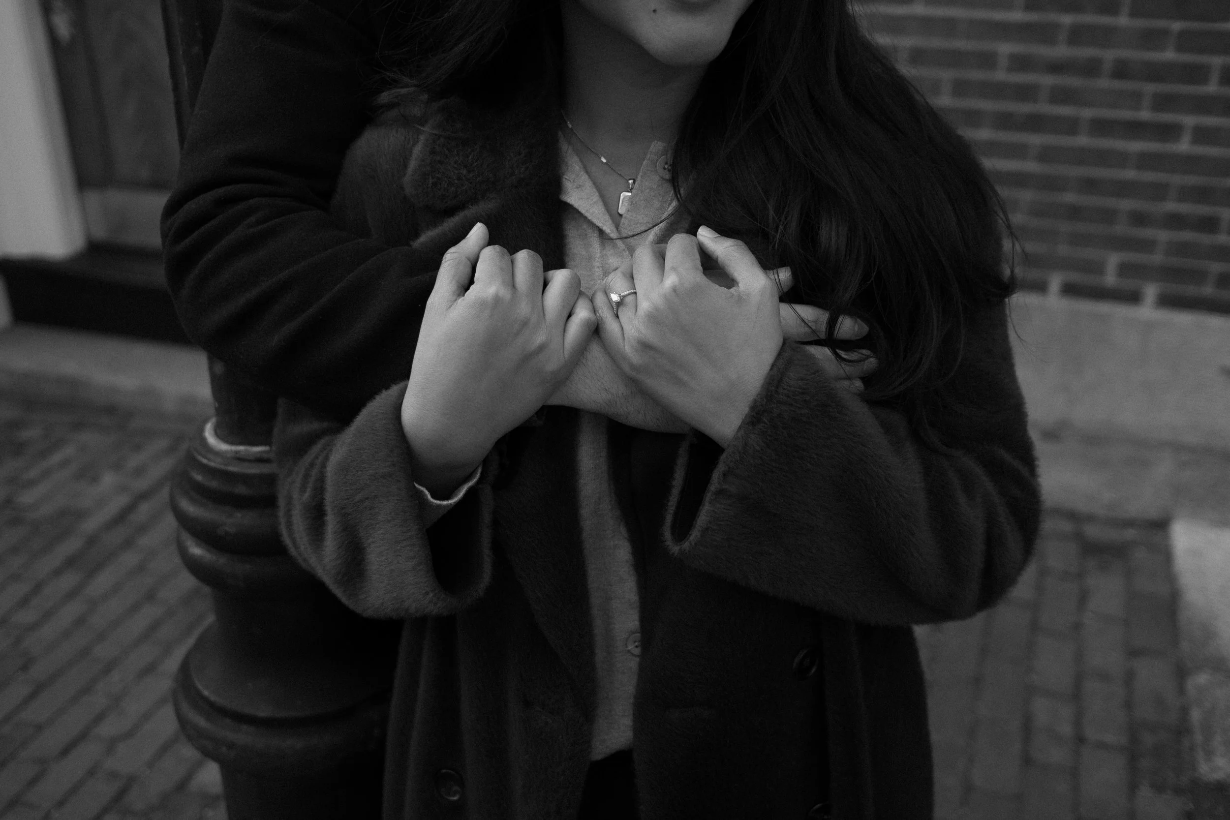 A woman wearing a dark coat and a necklace, holding her hands over her chest, standing against a brick wall.