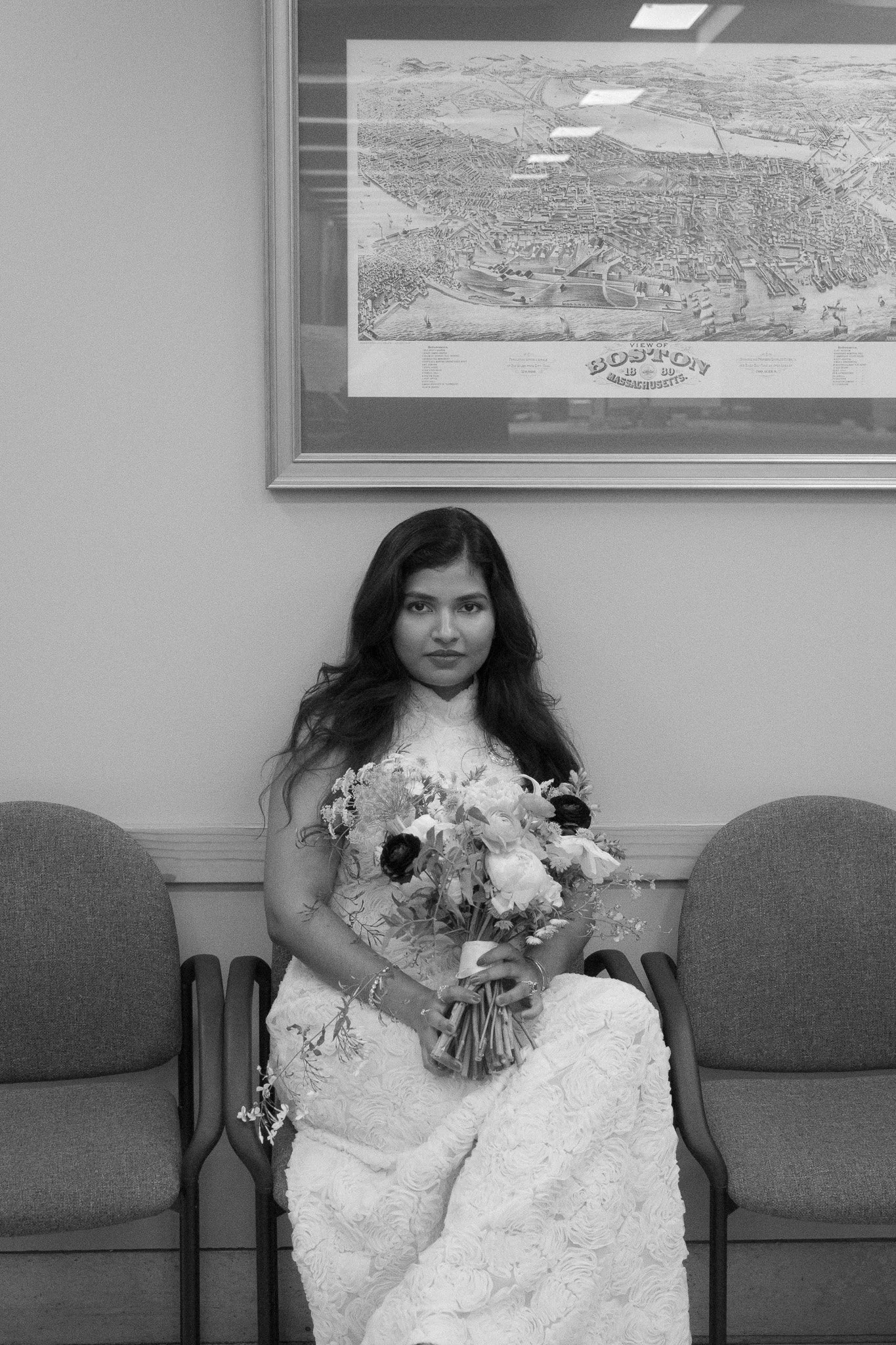 A young woman wearing a wedding dress sitting on a chair, holding a bouquet of flowers, in a waiting room with empty chairs and a large map of Boston, Massachusetts hanging on the wall behind her.