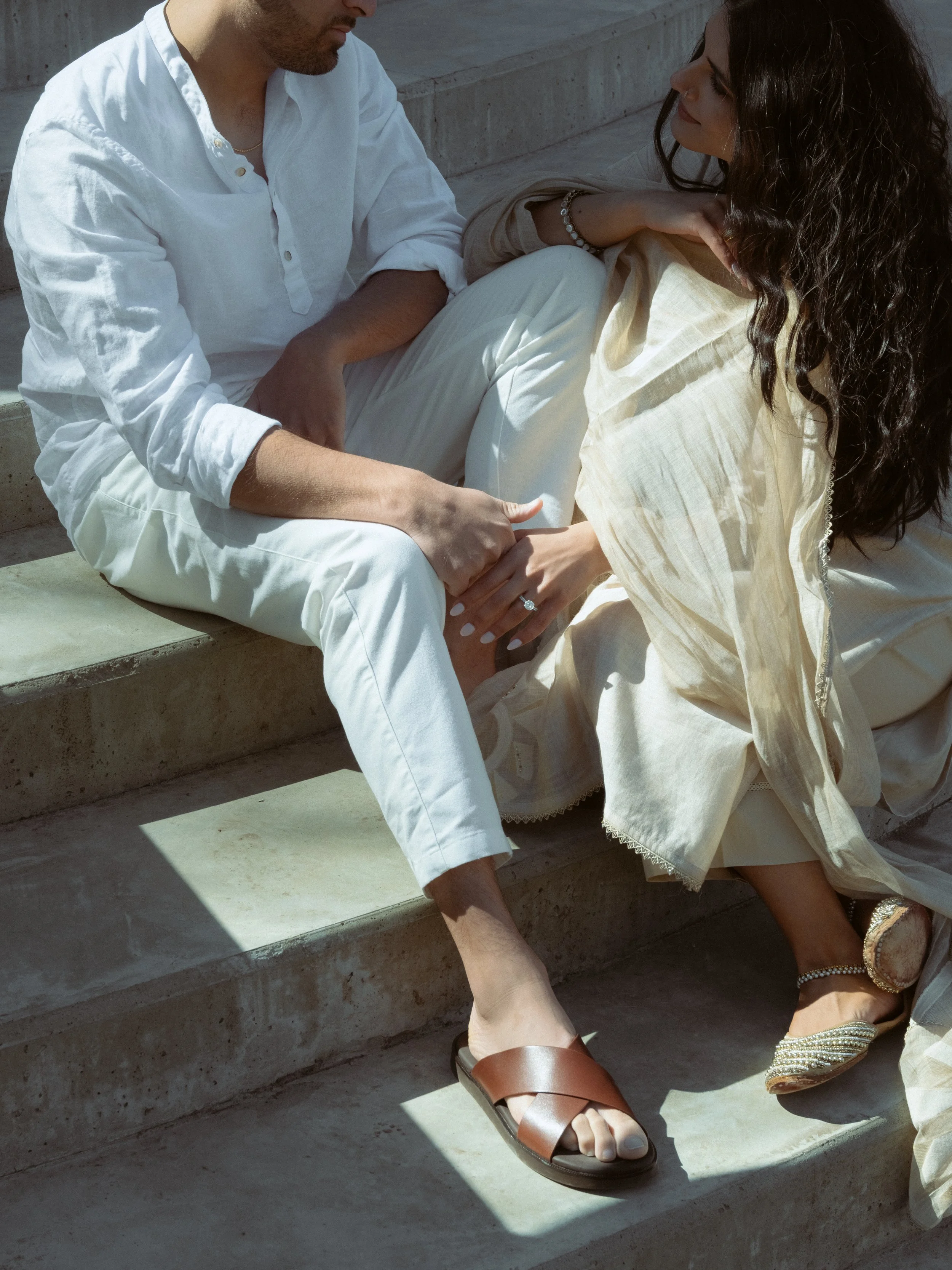Two people sitting on concrete stairs holding hands. One person is wearing brown sandals, and the other is wearing gold shoes. The person on the left is wearing white pants and a white shirt, while the person on the right is wearing cream-colored clothing and a pearl bracelet.