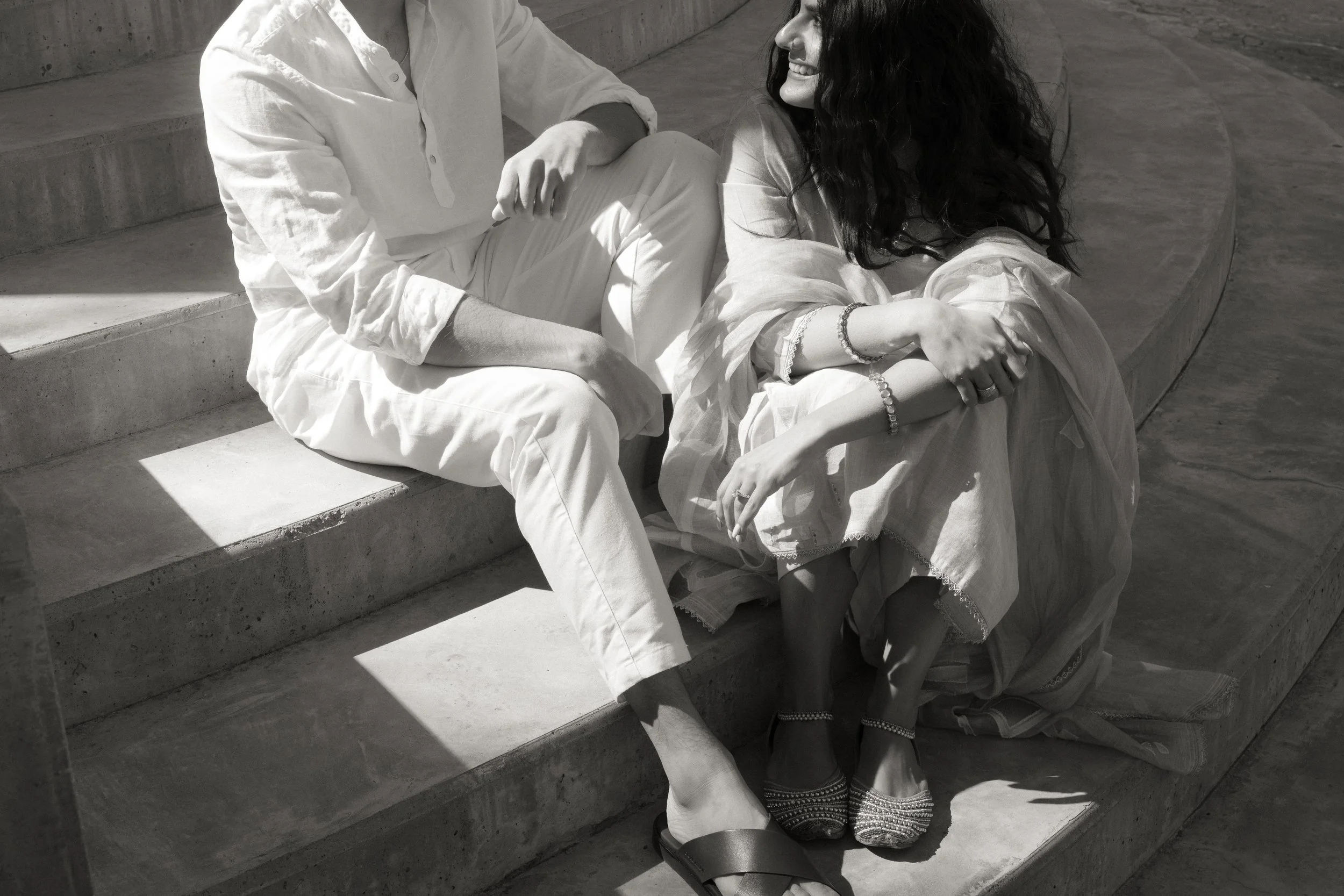 A black-and-white photo of a woman sitting on a concrete staircase, smiling at a man sitting near her. The woman has long curly hair and is dressed in traditional Indian clothing, wearing jewelry. The man is wearing light-colored clothing and a white shirt.