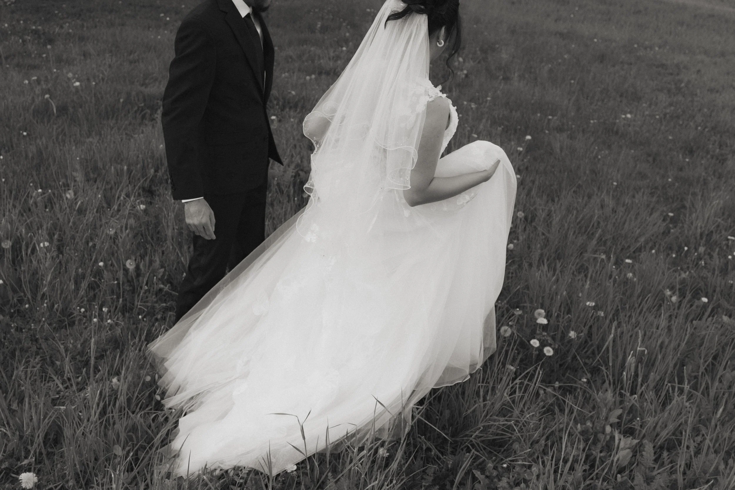 A bride in a wedding gown and veil standing in a grassy field near a groom dressed in a dark suit.