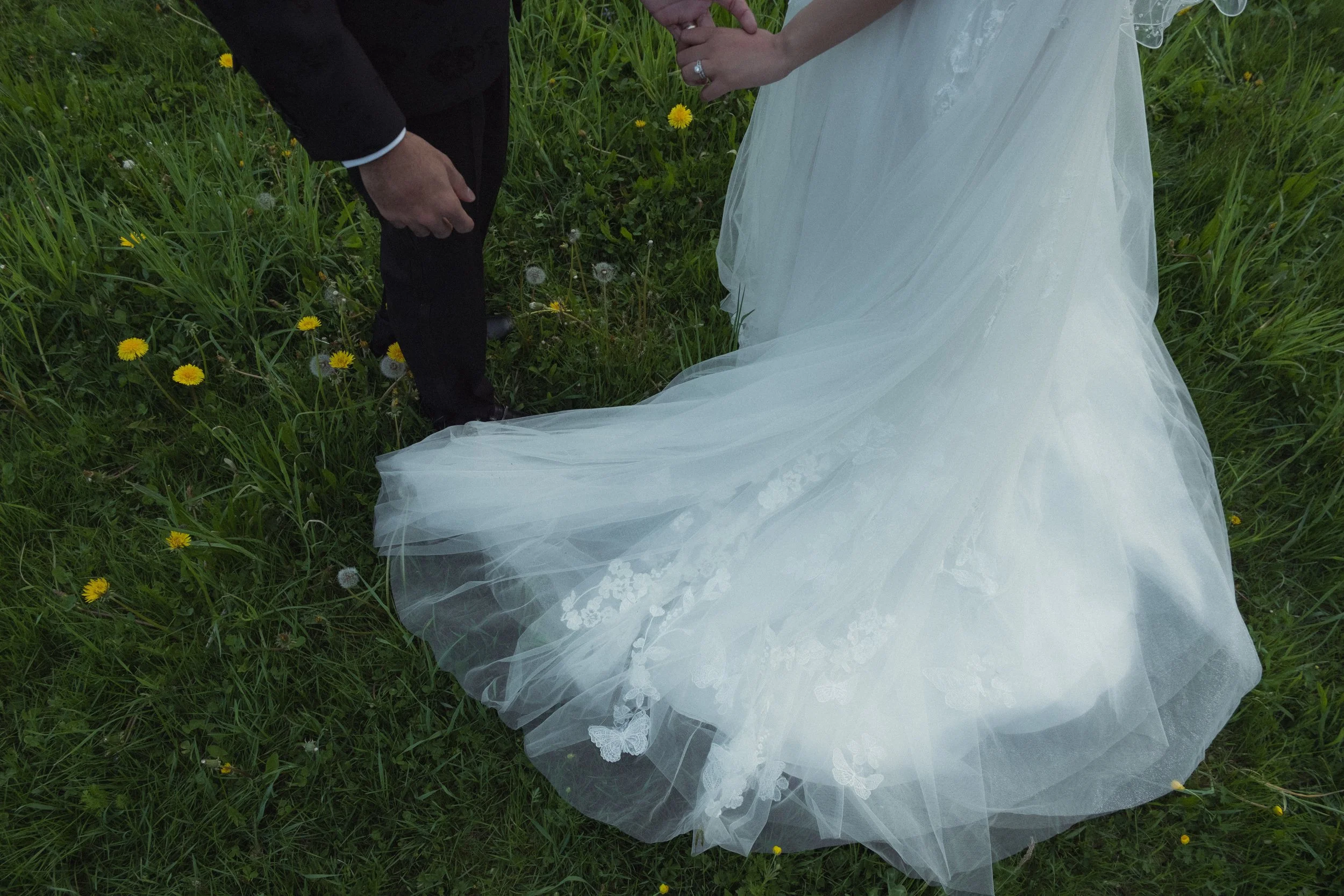 Close-up of a bride and groom holding hands while standing on grass with yellow flowers, the bride wearing a white dress and the groom wearing a black suit.