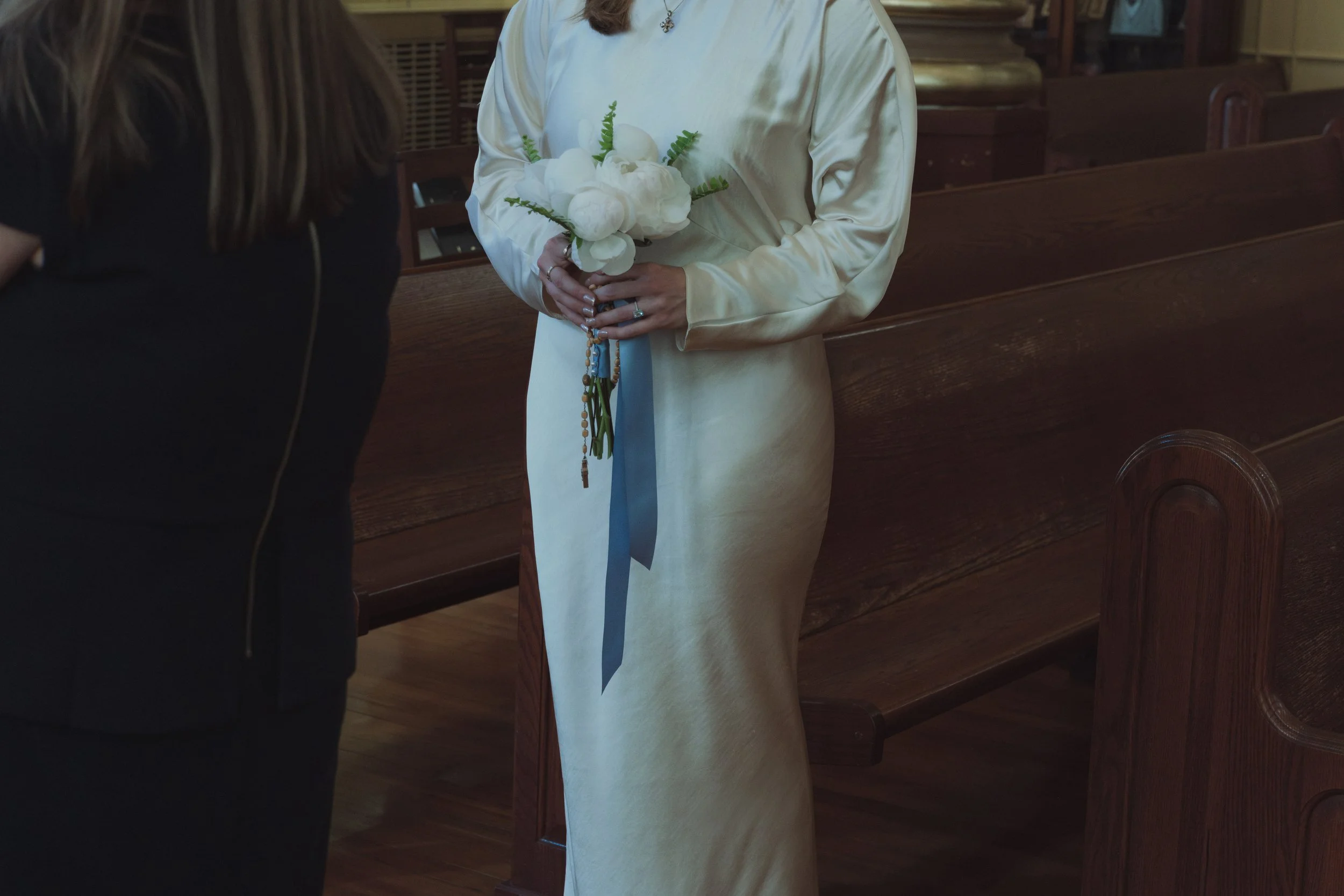 Person in a white satin dress holding a bouquet of white flowers and green ferns inside a church.