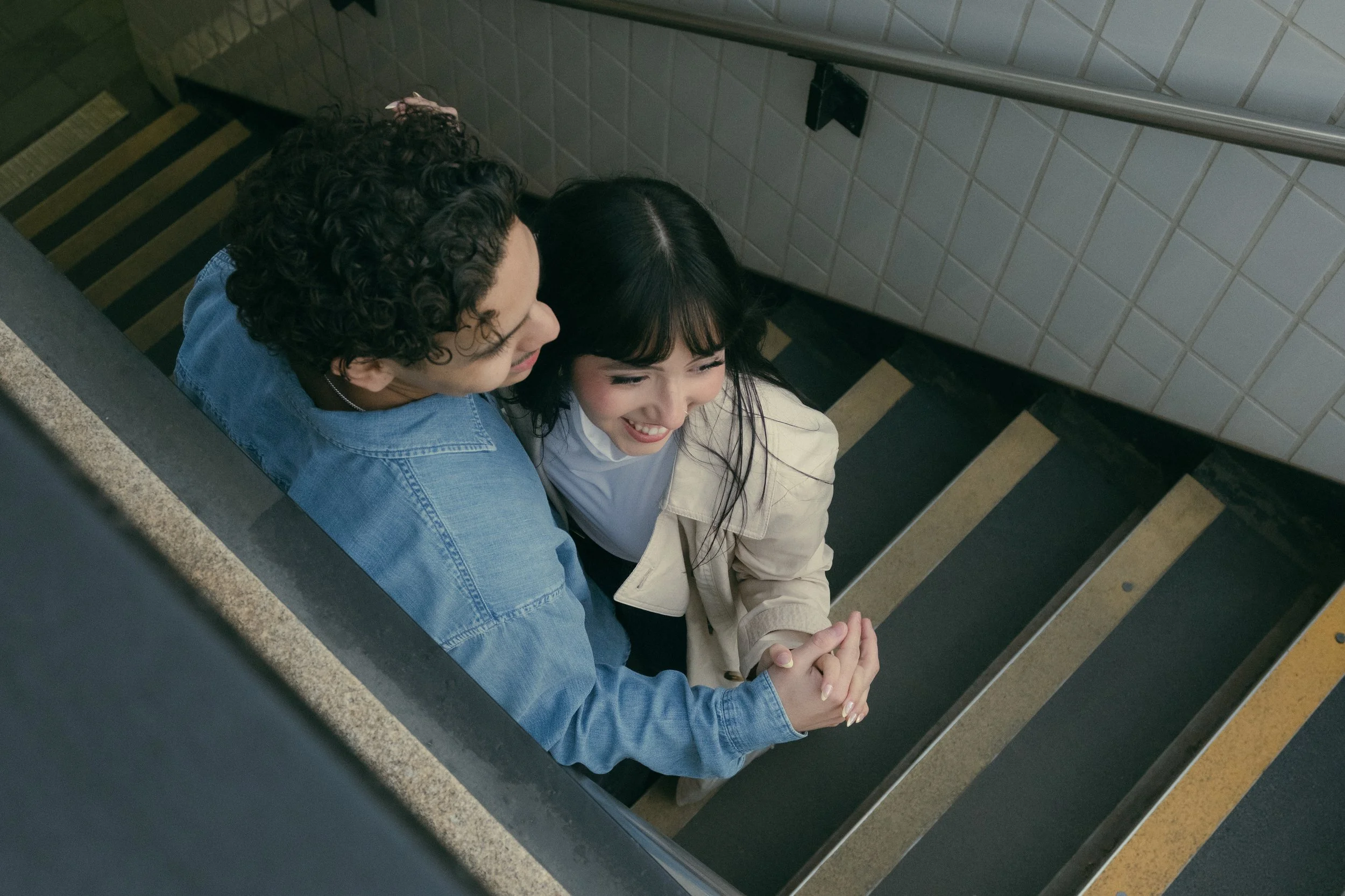 A young man with curly hair and glasses, and a young woman with dark hair and bangs, sitting close together on a staircase, holding hands and smiling. The staircase has black and yellow striped safety markings.