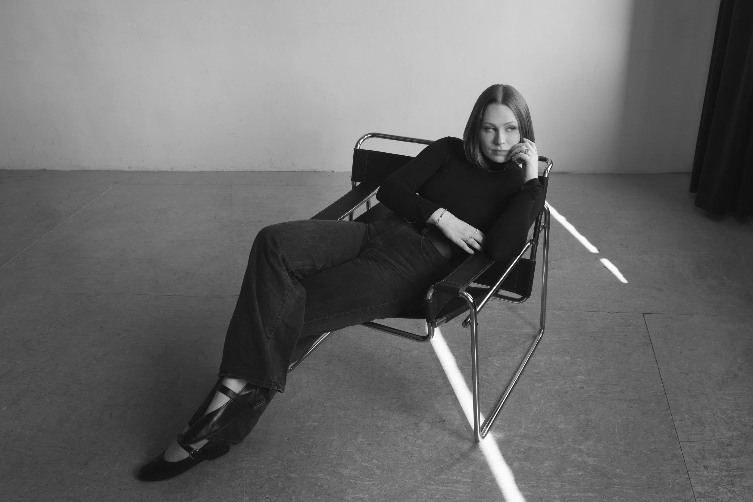 A woman sitting in a lounge chair with her legs crossed, resting her chin on her hand, looking to the side in a minimal indoor space with a plain wall and curtain, in black and white photograph.