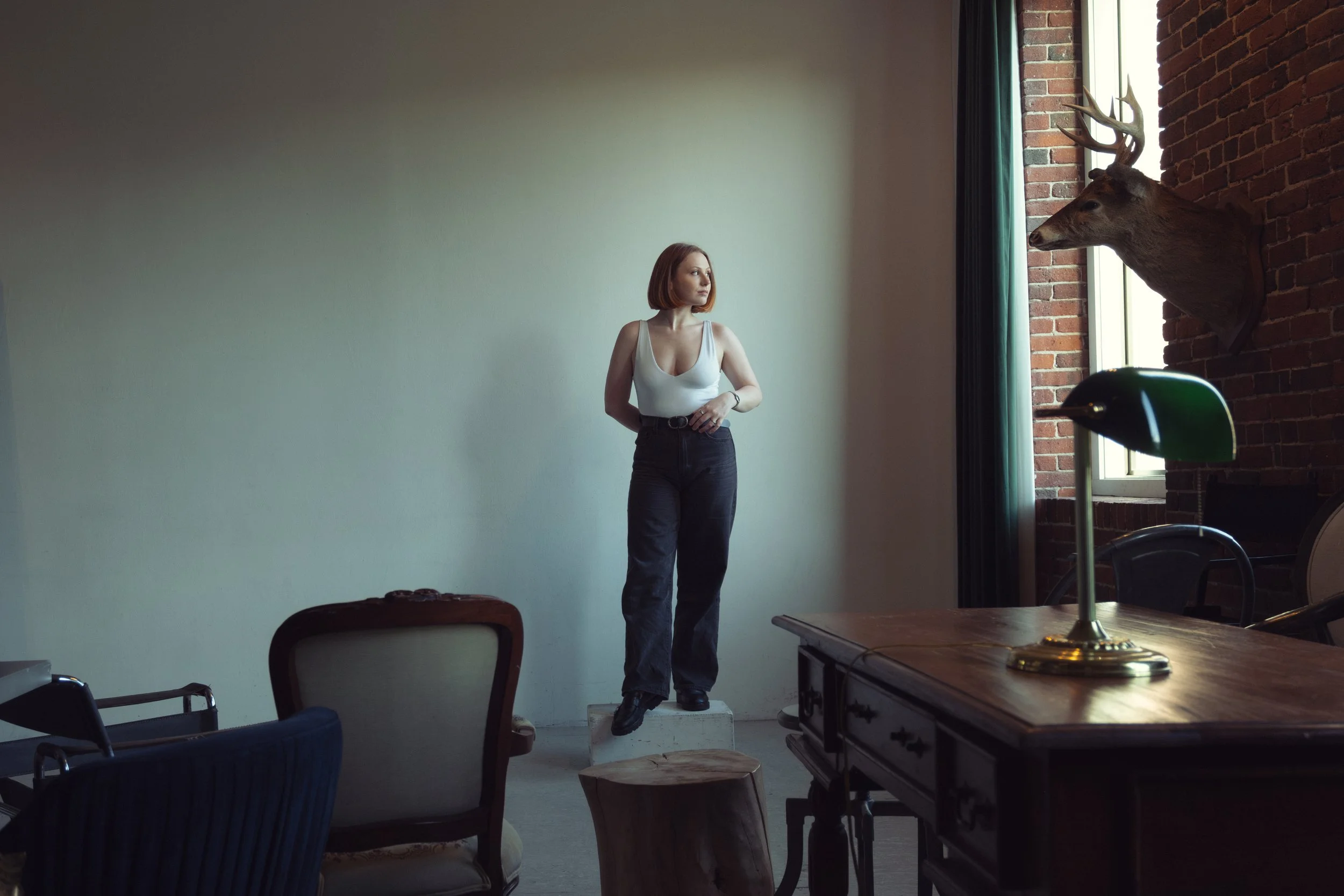 A woman with red hair wearing a white tank top and black pants standing on a small platform inside a room with brick wall and large window, looking at a mounted deer head on the brick wall.