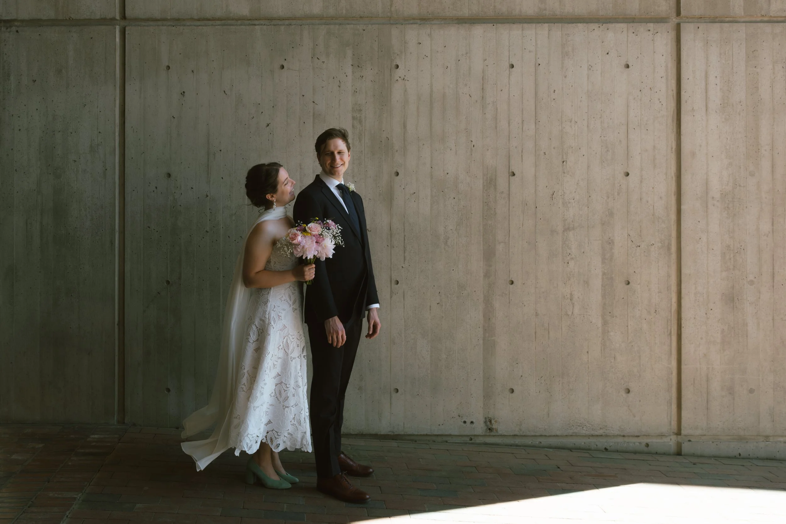 A bride and groom standing against a concrete wall, the bride holding a bouquet of pink and white flowers, both smiling, dressed in wedding attire.