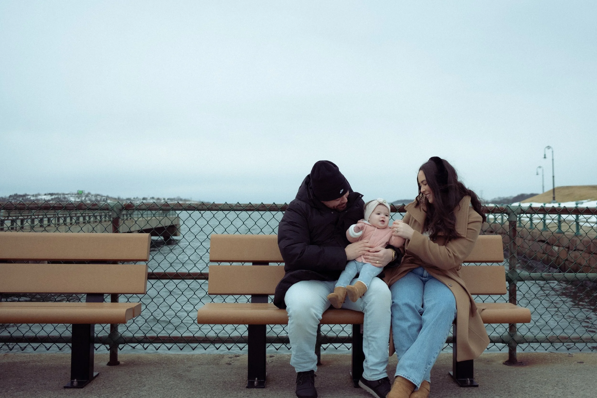 A family of three sitting on a park bench near a body of water. The man is holding a baby girl, and the woman is smiling at them, all dressed in warm clothes.