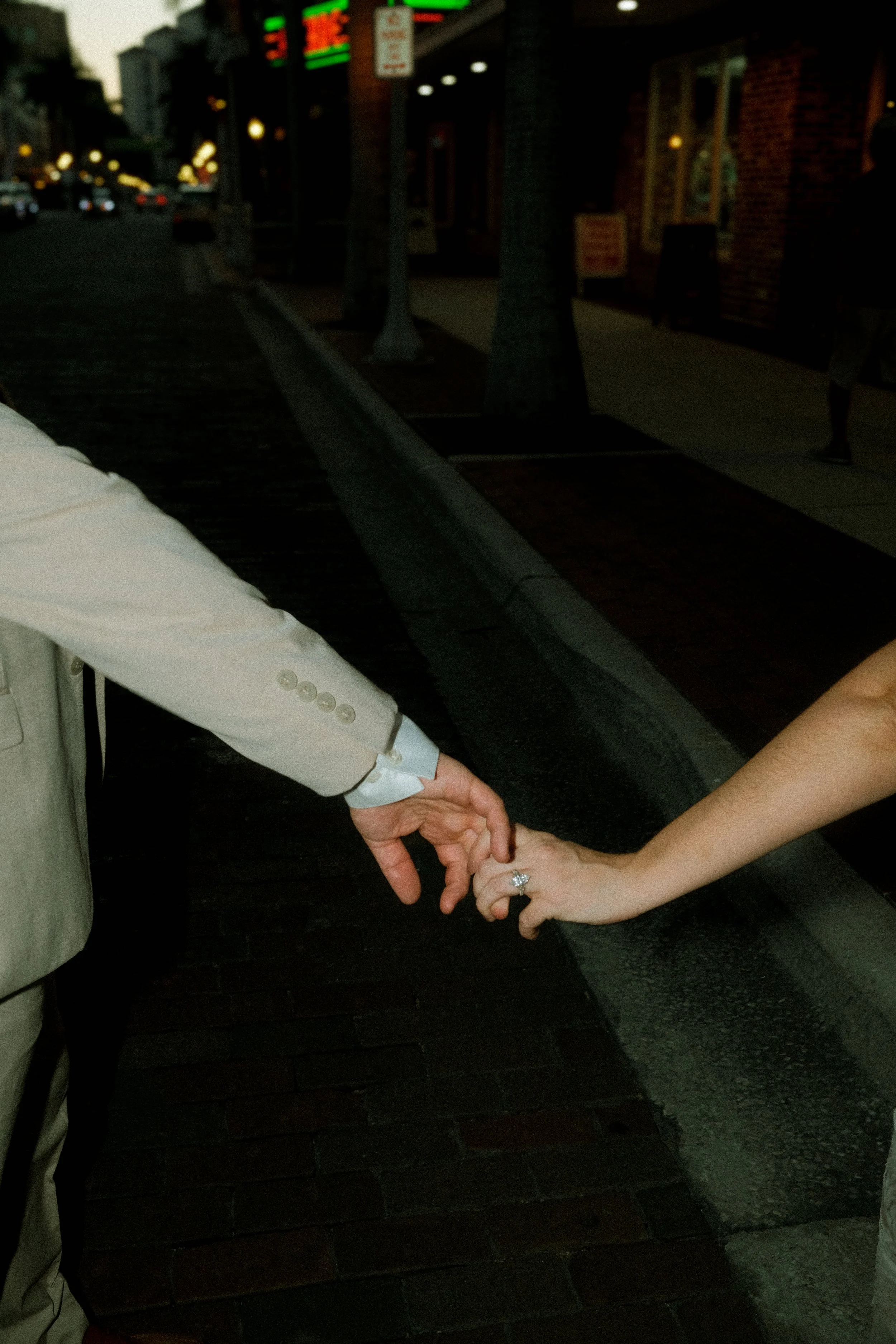 A couple holding hands, with the man on the left wearing a light-colored suit and the woman on the right showing an engagement ring, on a city sidewalk at night.
