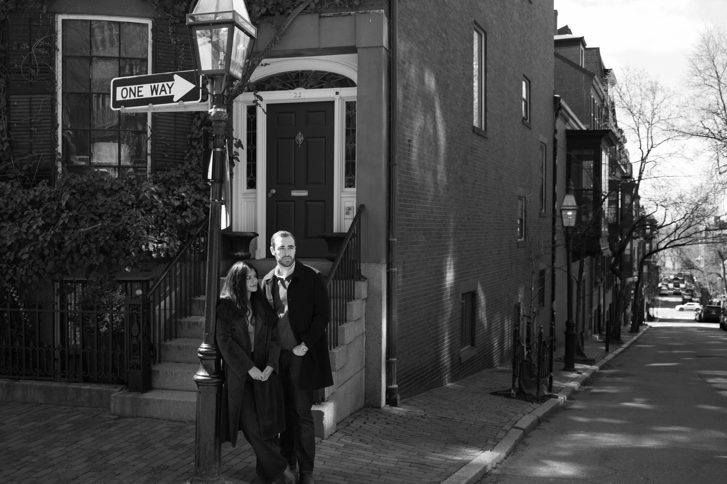 A black and white street scene with a man and woman standing near a lamppost, in front of a building entrance with stairs. The street has parked cars and leafless trees.