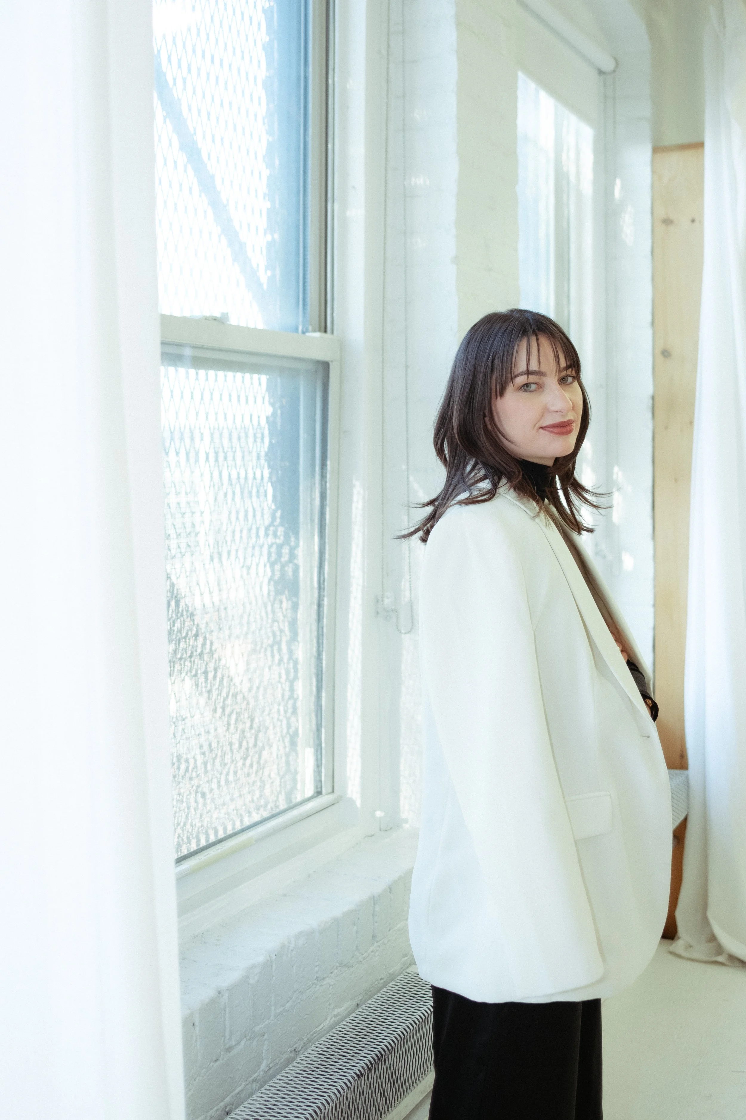 A woman with dark hair and bangs, wearing a light-colored blazer and dark pants, standing next to a window with frosted glass, in a bright room with white walls and curtains.