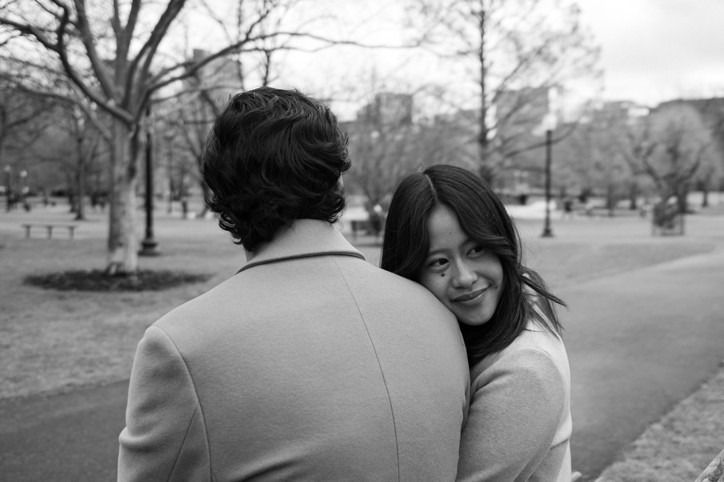 A black and white photo of a woman smiling while hugging a man, both standing outdoors in a park with trees and benches.