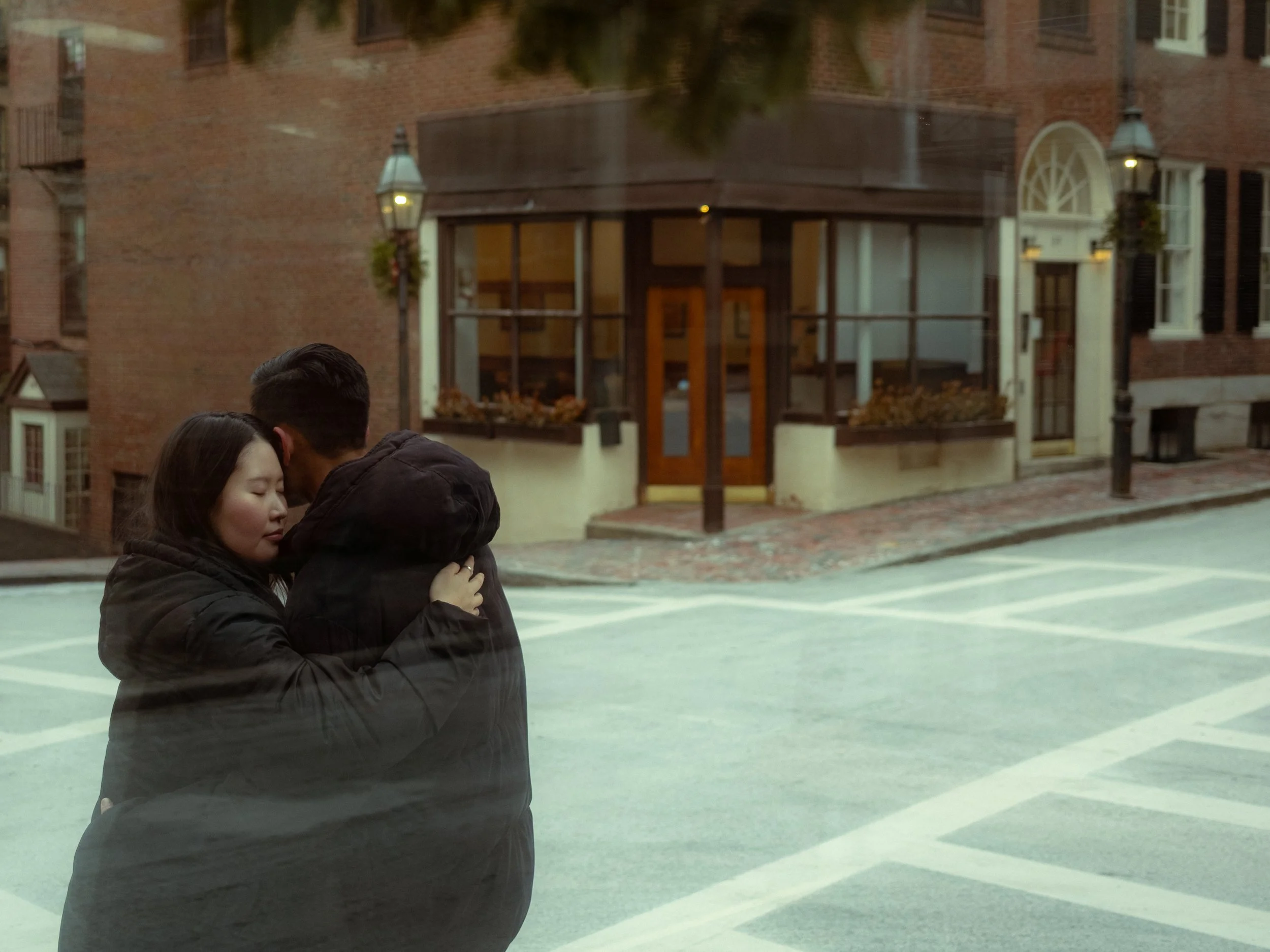 A couple embraces on a city street with brick buildings and street lamps in the background.