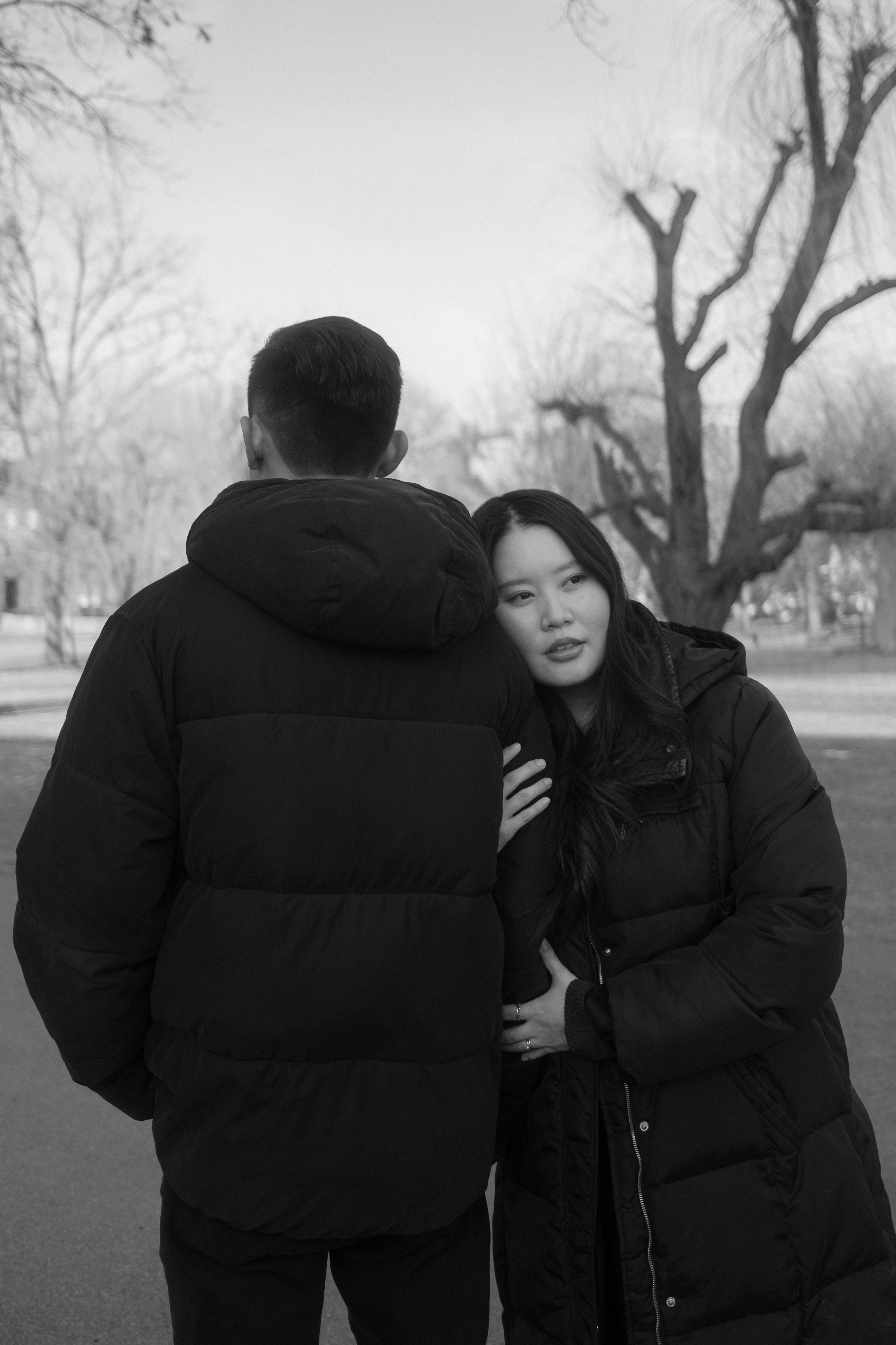 A black and white photo of a man and a woman standing outdoors in winter, dressed in puffer jackets. The woman rests her head on the man's shoulder, and they appear to be in a park with bare trees in the background.