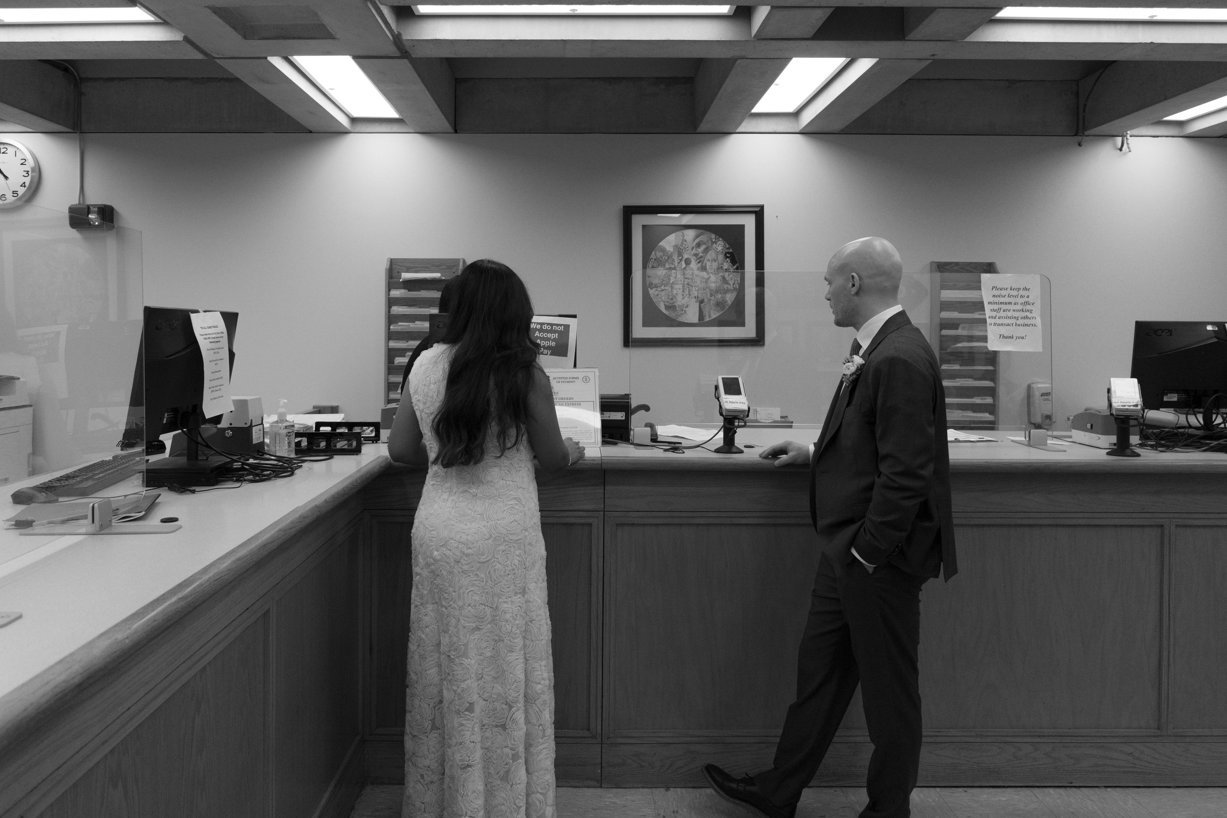 A woman and a man standing at a bank counter, engaging in a transaction or conversation. The woman is facing the counter with her back to the camera, and the man is on the customer side, looking at her. The setting appears to be a bank or financial institution with computers, signs, and a partition on the counter.