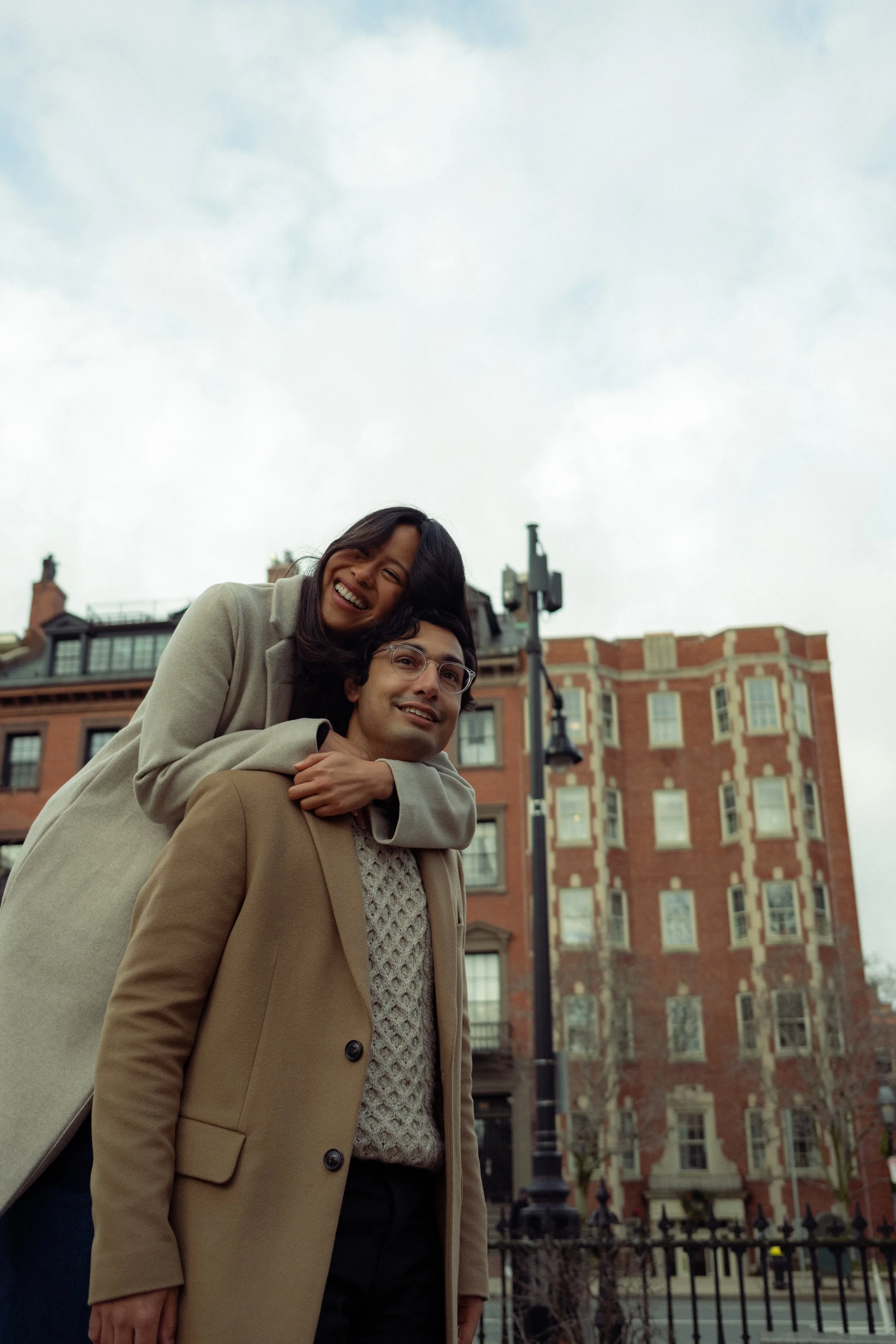 A woman with dark hair and a beige coat smiling and hugging a man with glasses and a beige coat outdoors in front of a red brick building.
