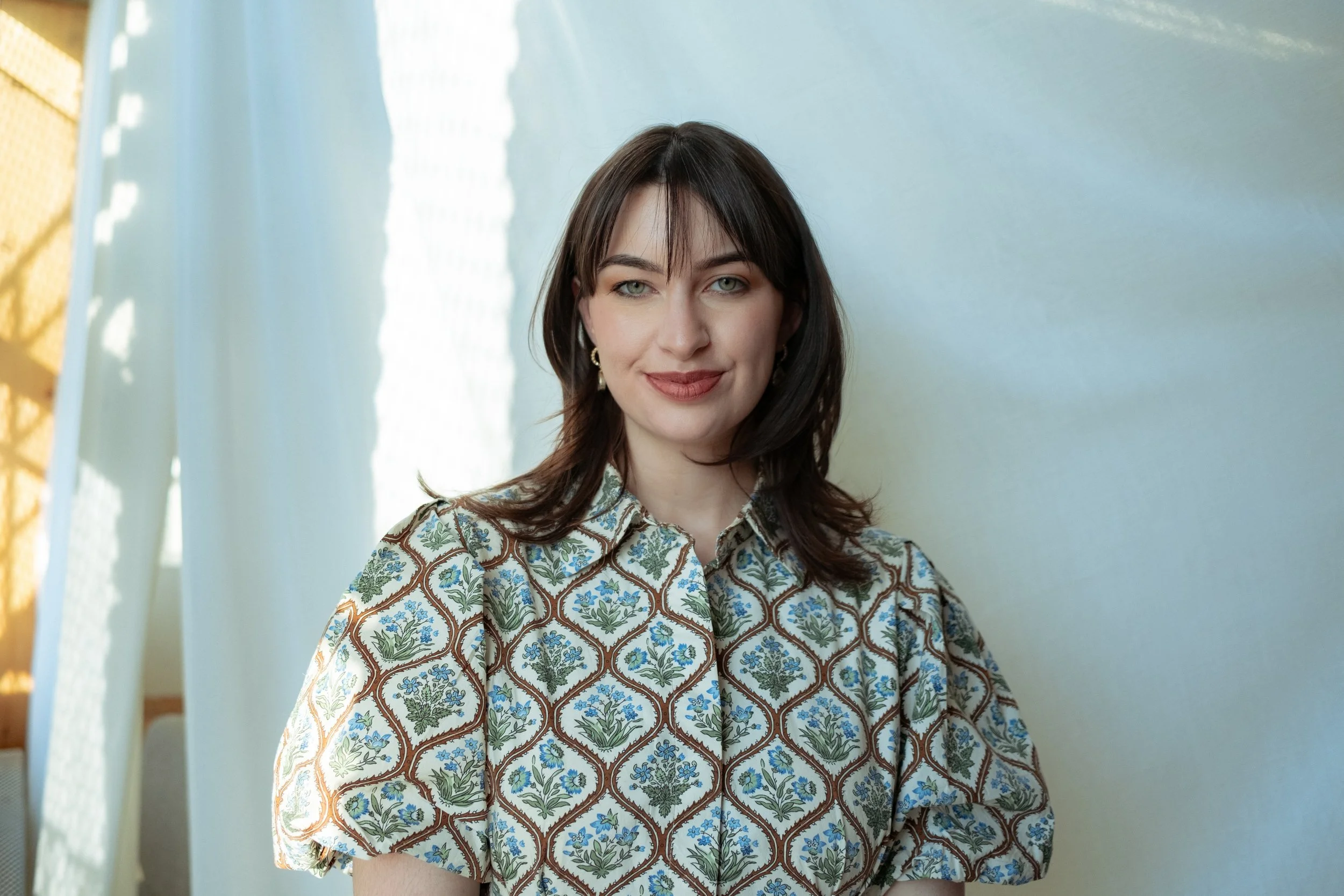 A young woman with dark hair and wearing a floral patterned blouse, standing indoors with a light-colored wall and bright window light in the background.