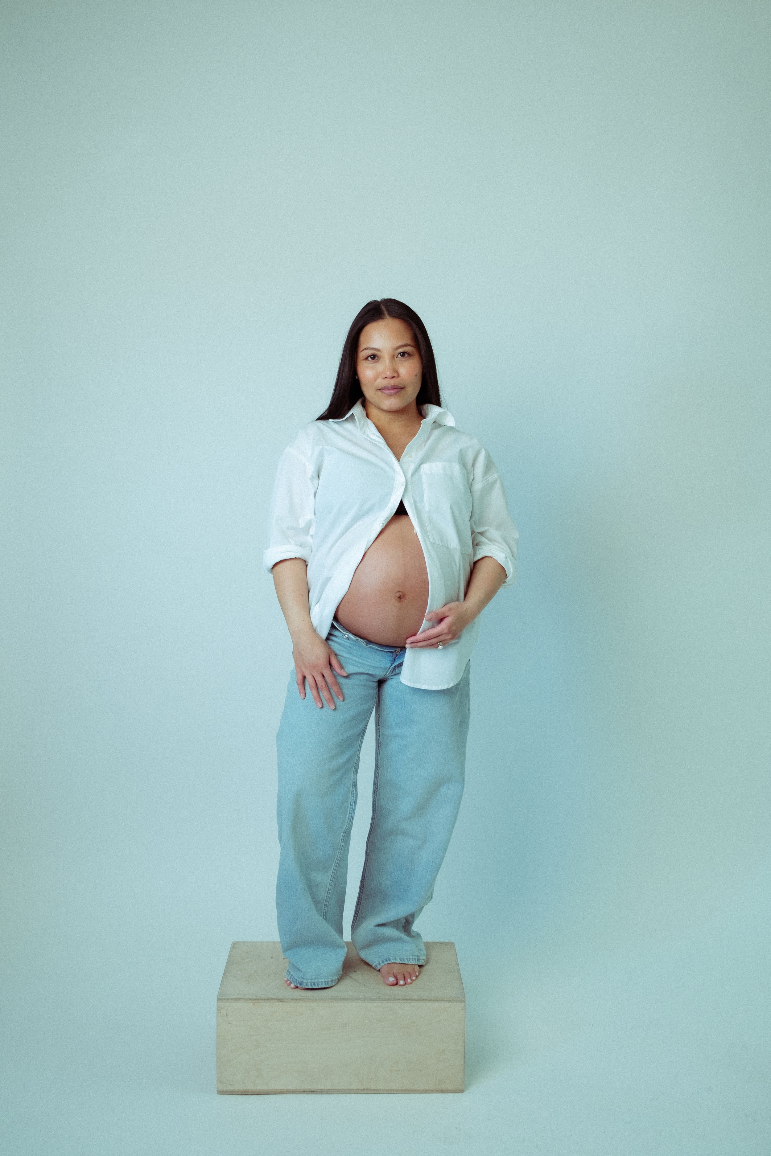 A pregnant woman standing barefoot on a wooden box, wearing an open white shirt and loose jeans, with her hand on her belly, in front of a plain light background.