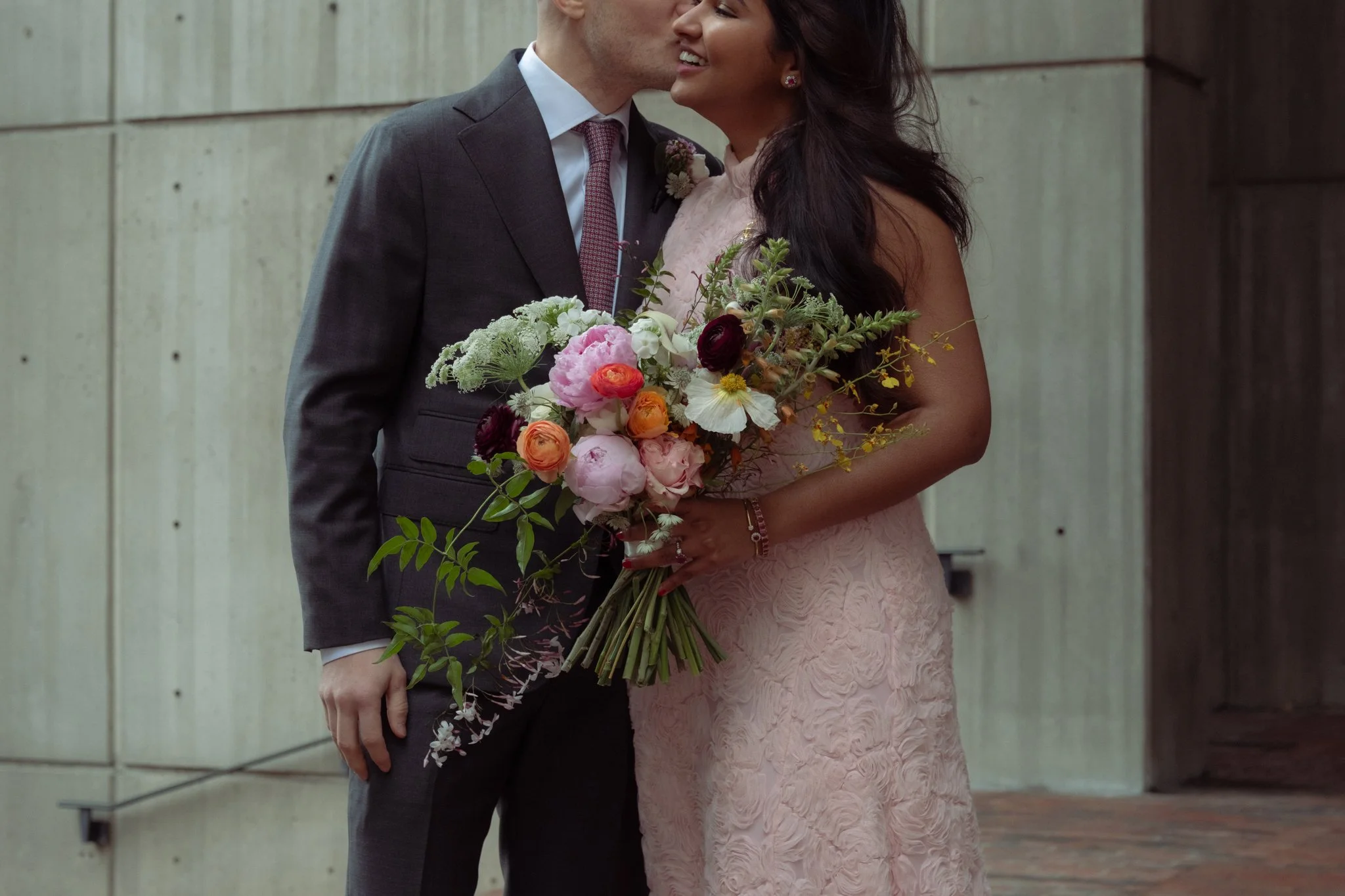 A couple, dressed formally, sharing a kiss at an outdoor event, with the woman holding a large bouquet of colorful flowers.