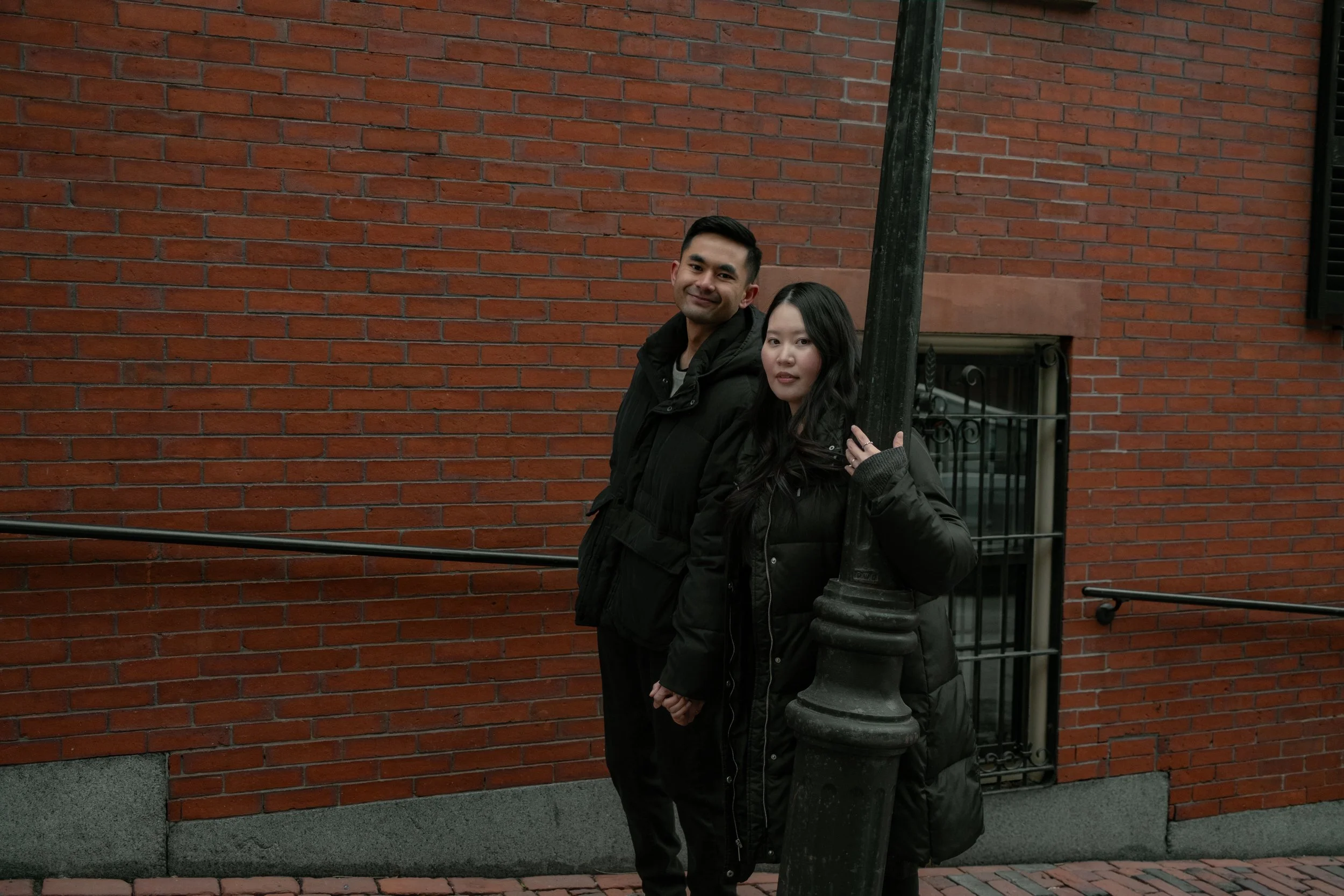 A man and woman standing on a brick sidewalk beside a red brick wall, holding hands, with the woman partially behind a black streetlamp, both wearing black jackets.
